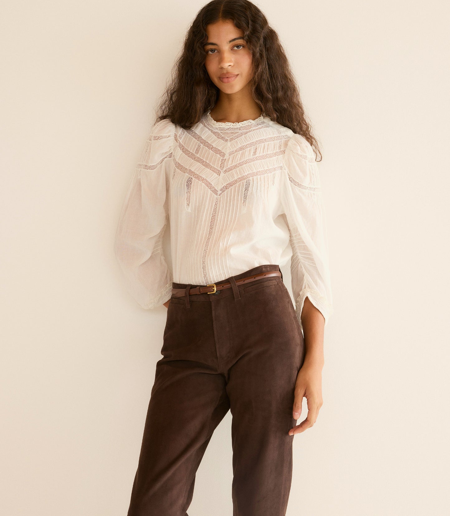 Woman wearing a white blouse with intricate patterns and brown pants against a beige background