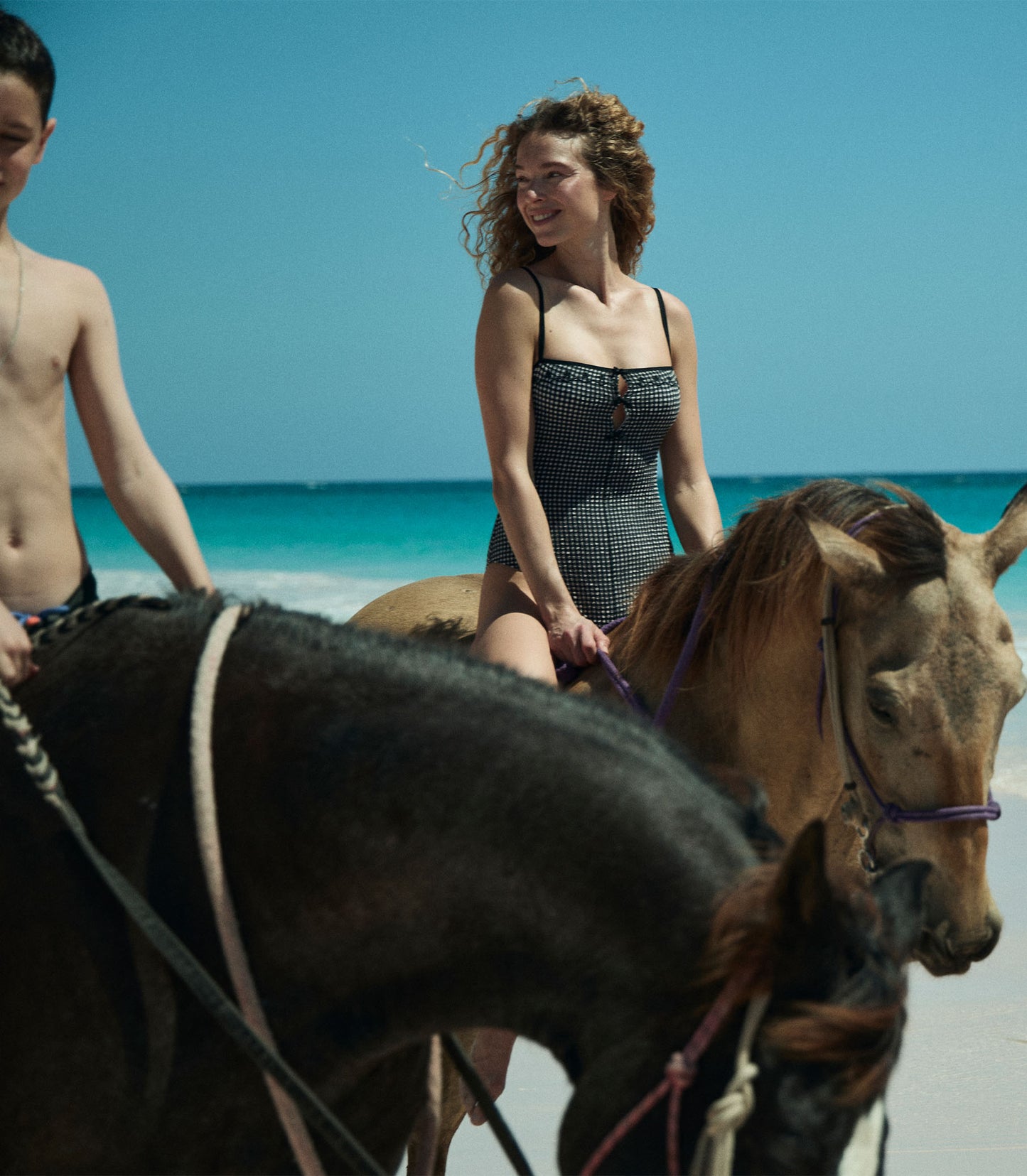 Heloise is wearing a black and white gingham one piece swimsuit while riding a horse on the beach.