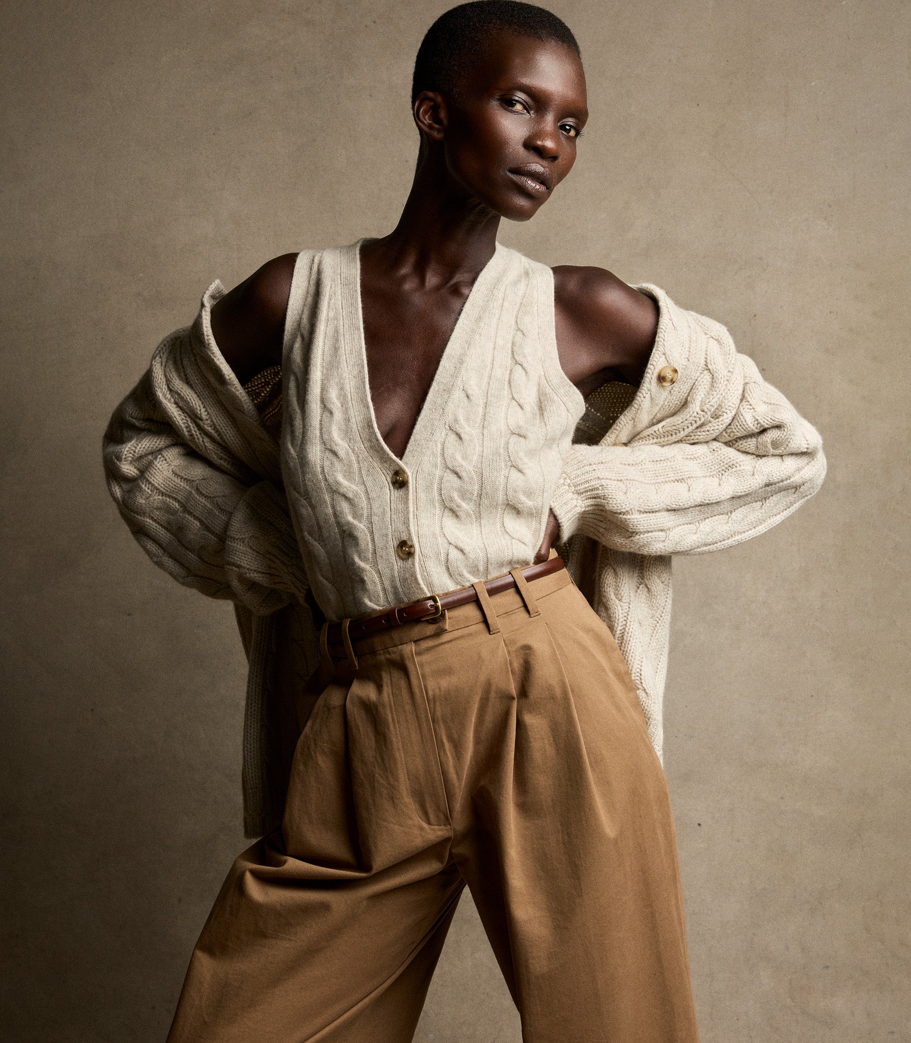 Person wearing a white cable knit vest and brown pants against a beige background