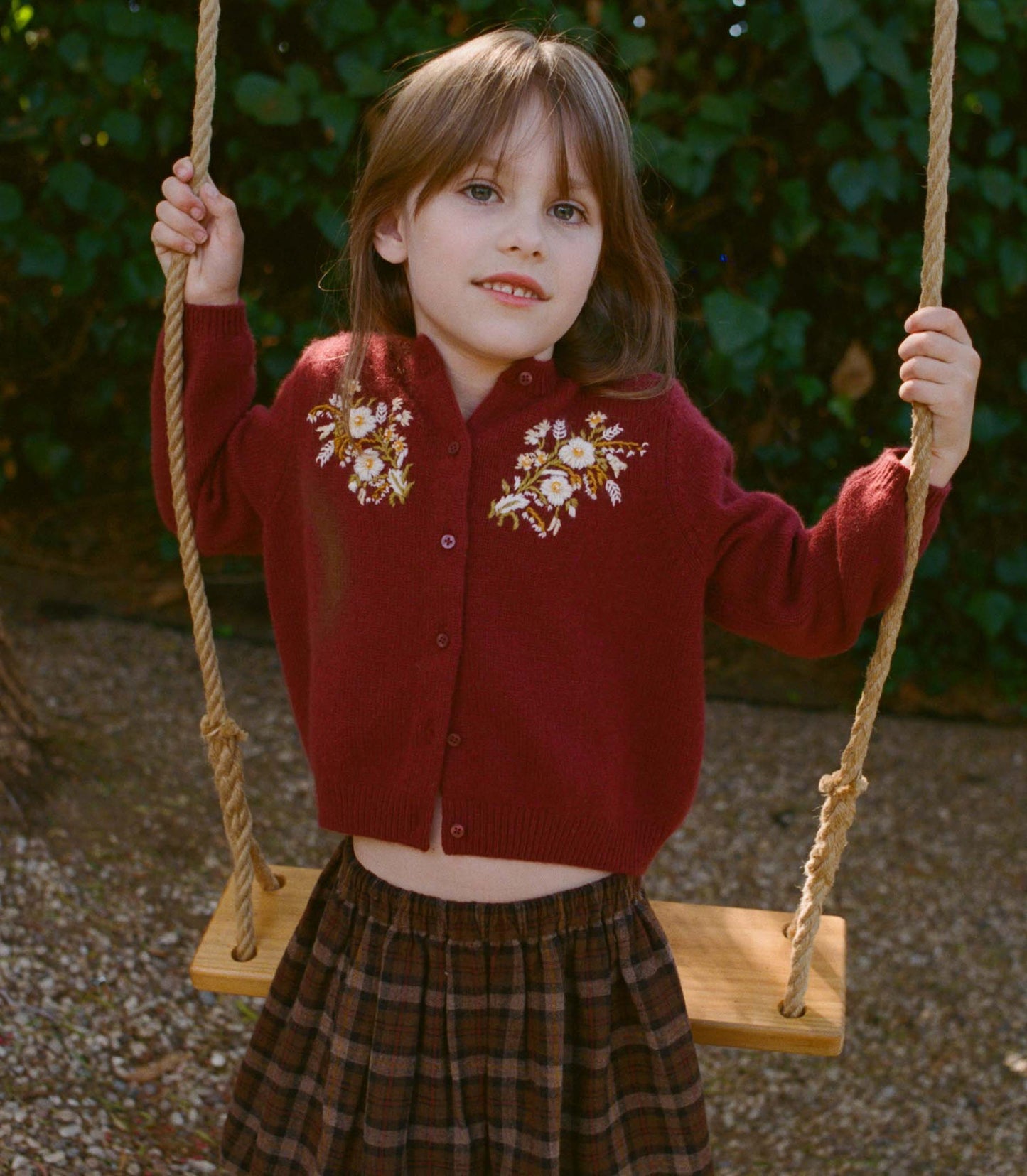Young girl in a red cardigan with floral patterns on a swing outdoors.