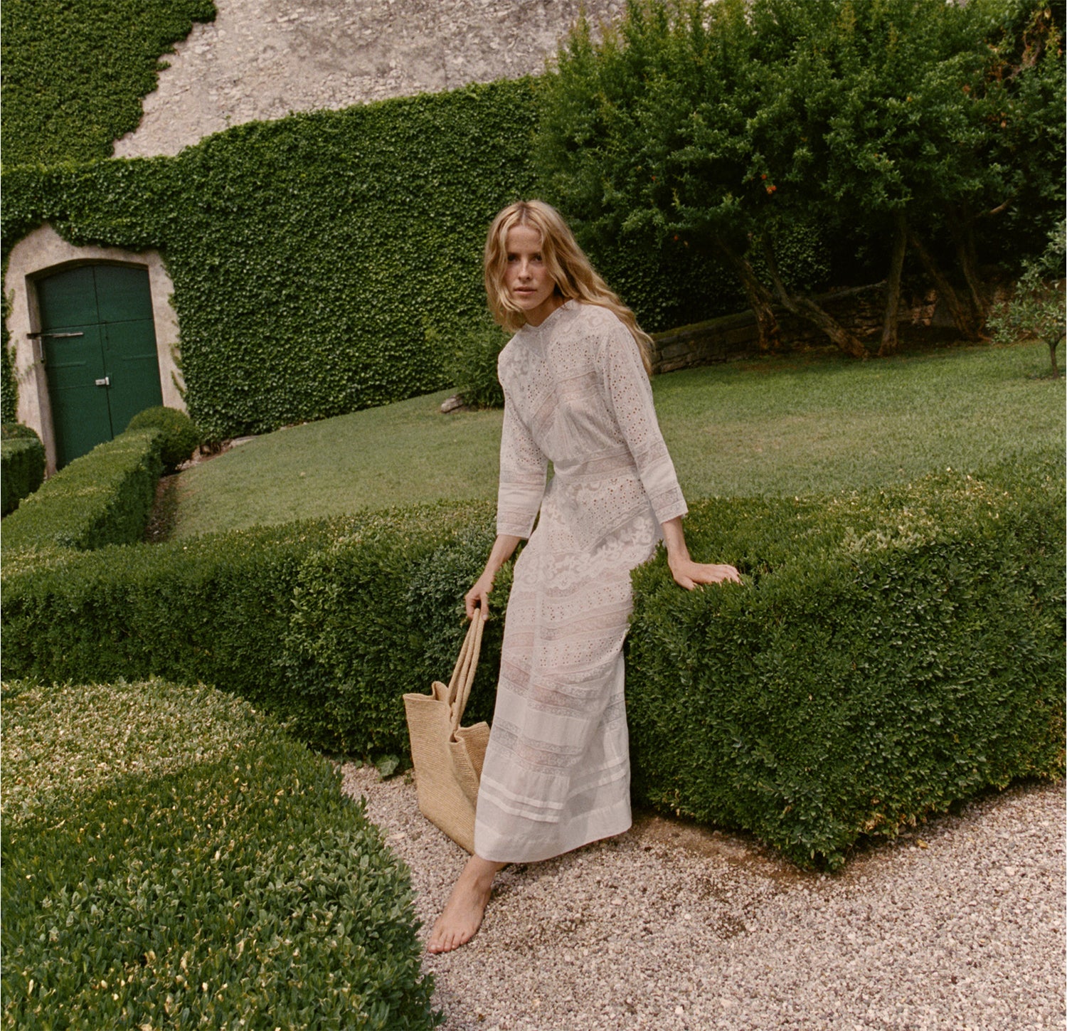 Woman in a light dress standing in a garden with manicured hedges and a green door.