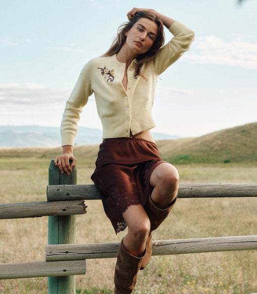 Woman sitting on a wooden fence in a field wearing a beige cardigan and brown skirt.