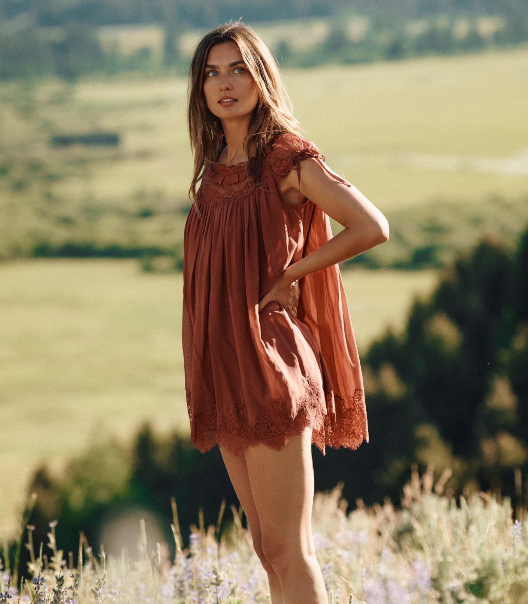 Woman in a rust-colored lace dress standing in a field with a scenic background