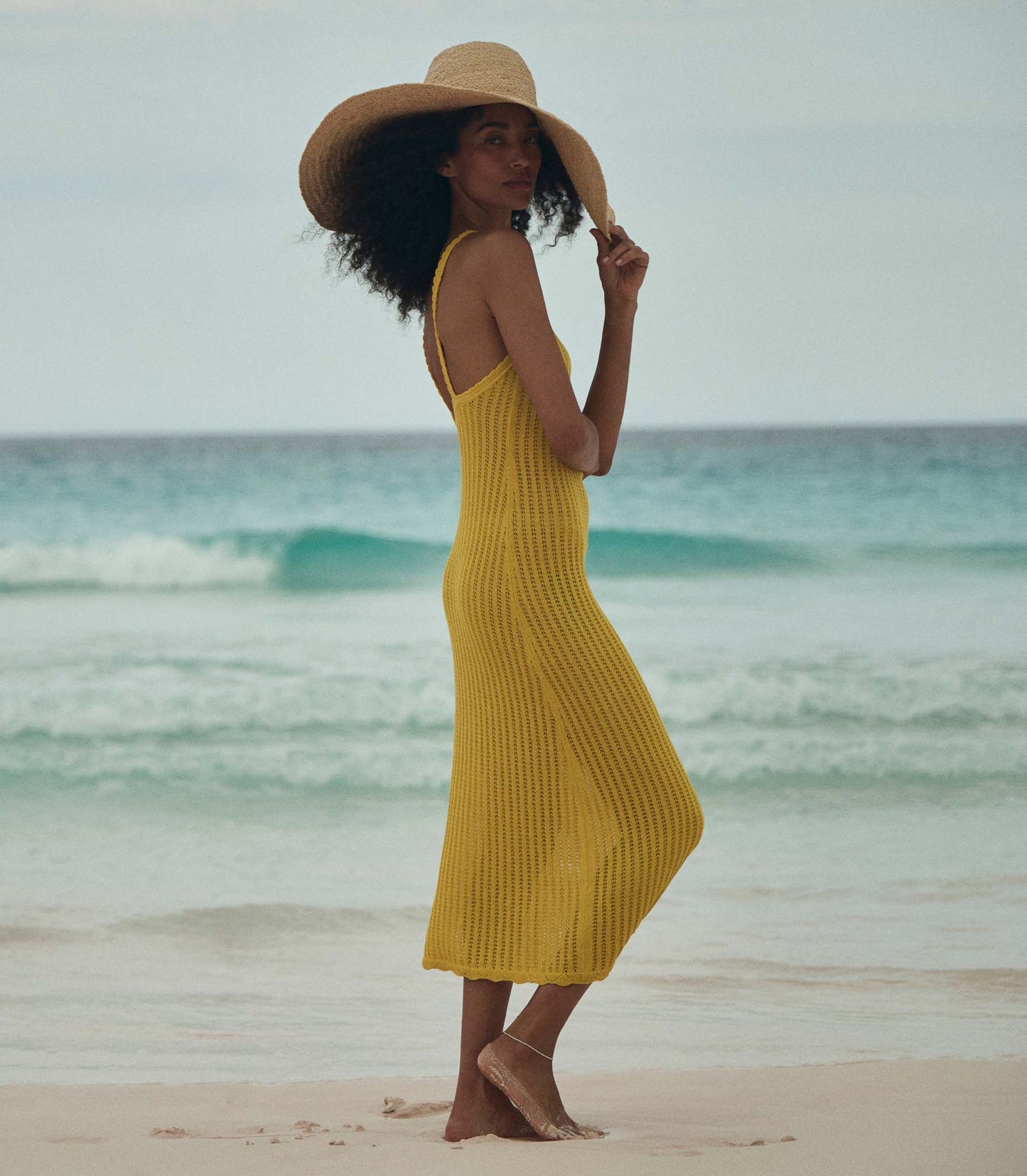Anais is wearing a sleeveless yellow knit maxi dress with a sun hat on the beach.