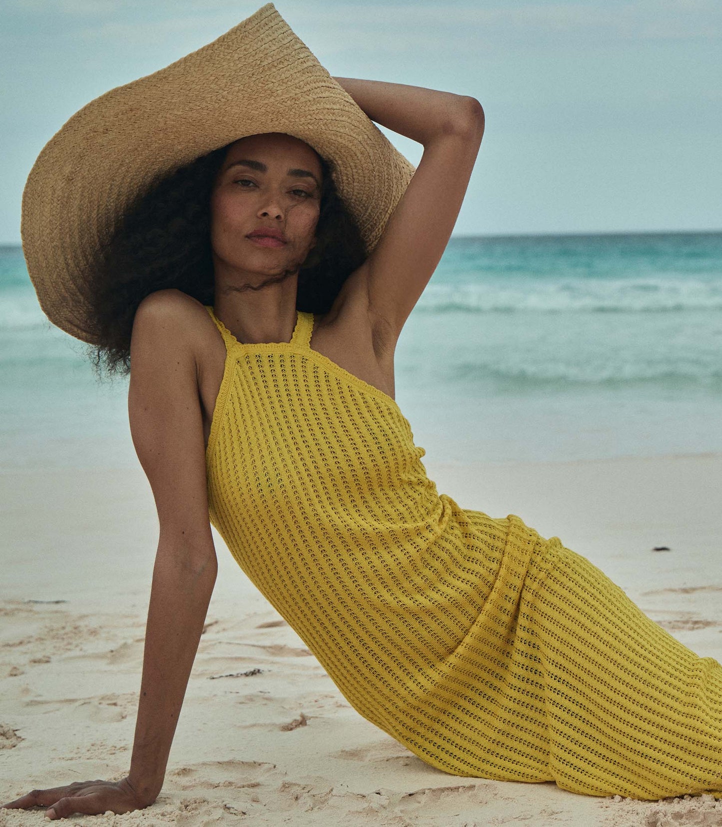 Anais is wearing a sleeveless yellow knit maxi dress with a sun hat on the beach.