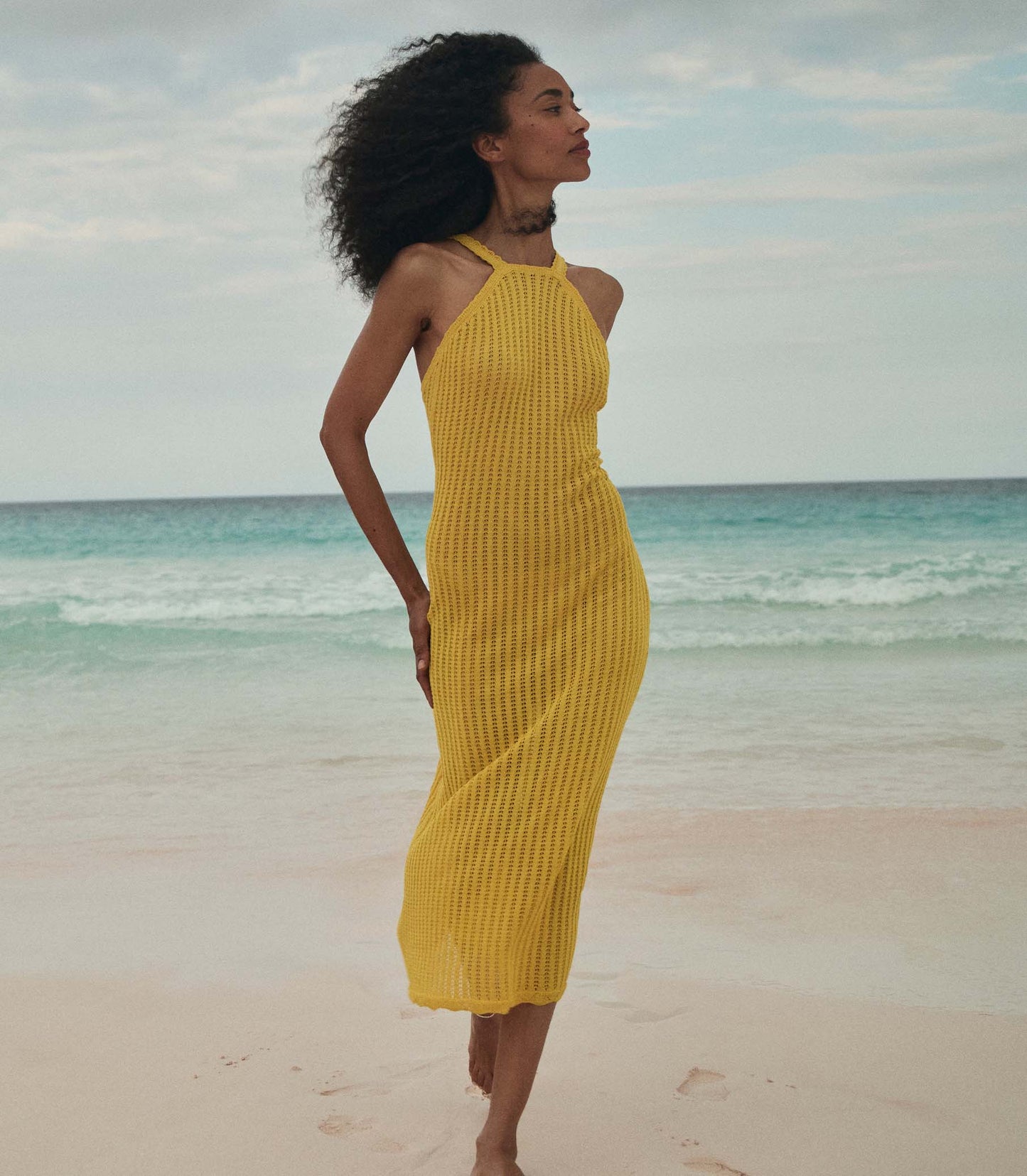Anais is wearing a sleeveless yellow knit maxi dress with a sun hat on the beach.