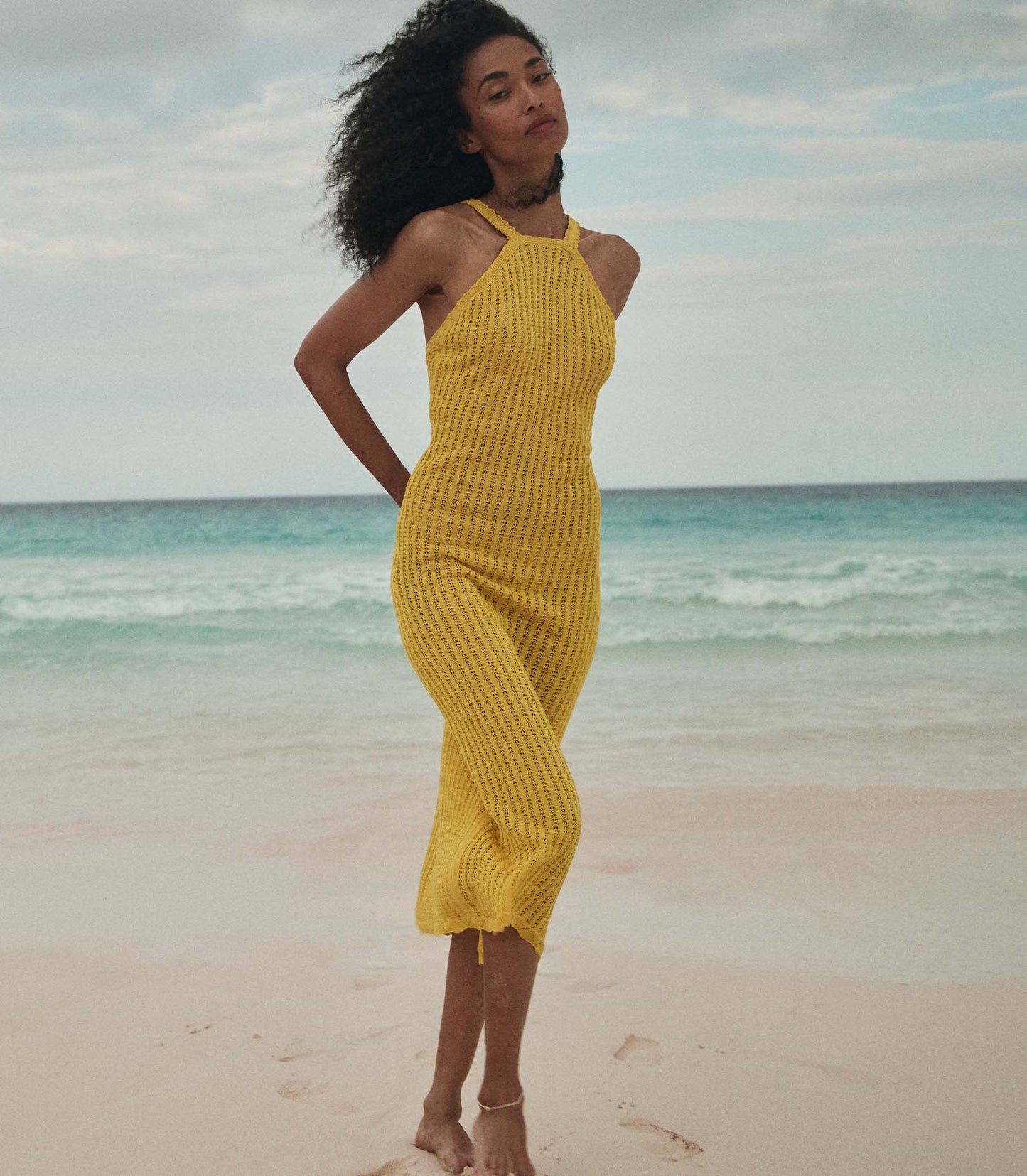 Anais is wearing a sleeveless yellow knit maxi dress with a sun hat on the beach.