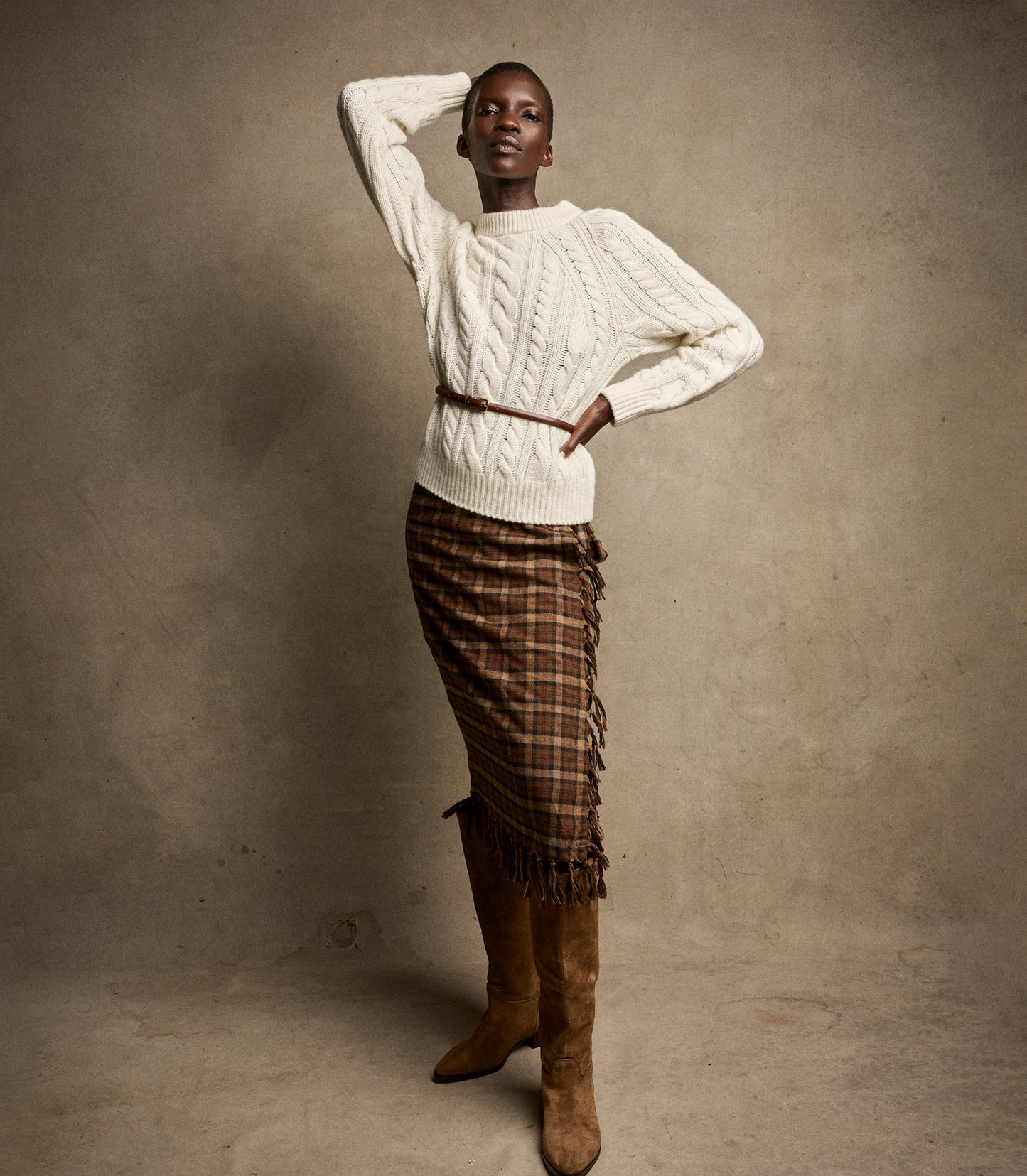 Woman wearing a white cable knit sweater and plaid skirt with brown knee-high boots against a beige background
