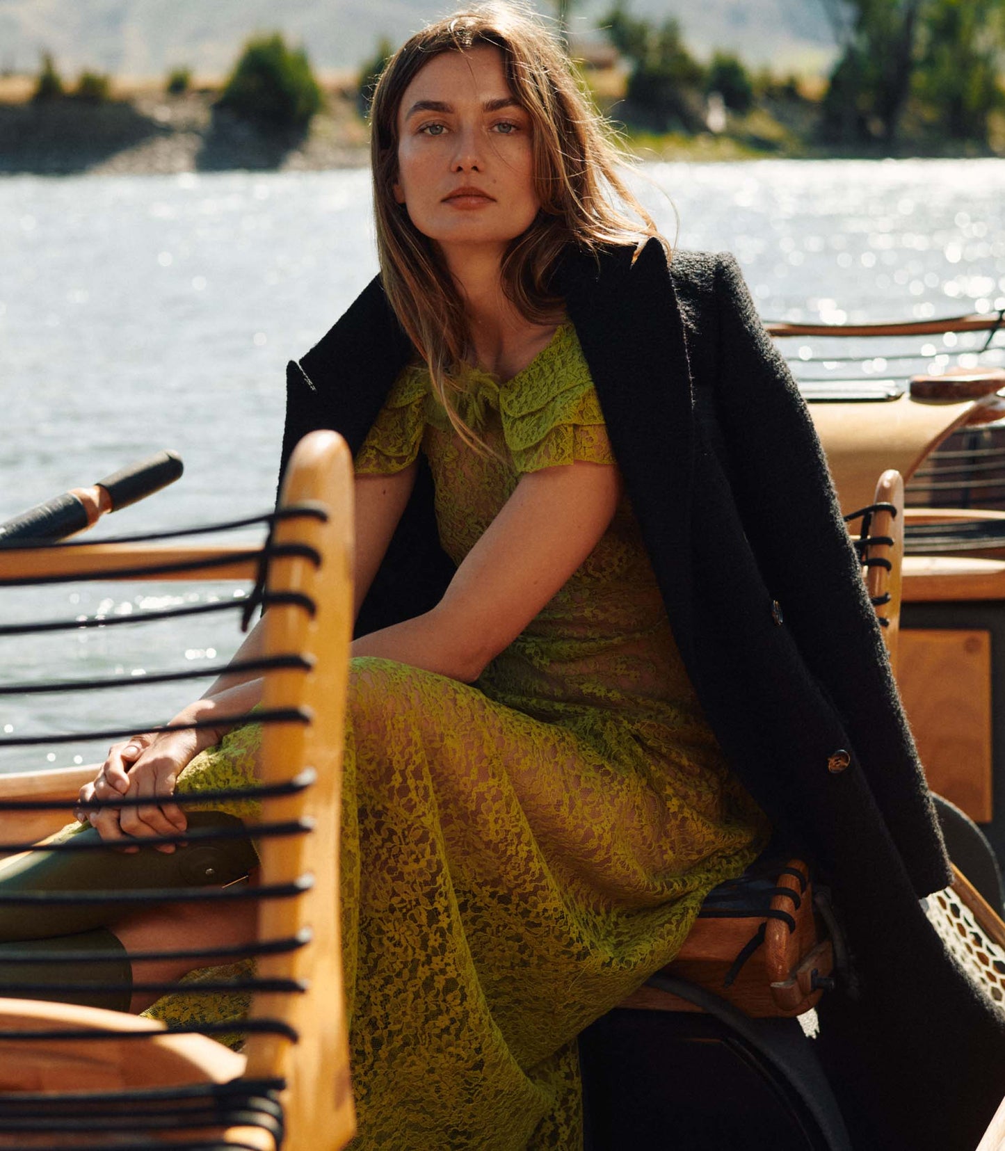 Woman in a green lace dress and black coat sitting outdoors by a body of water.
