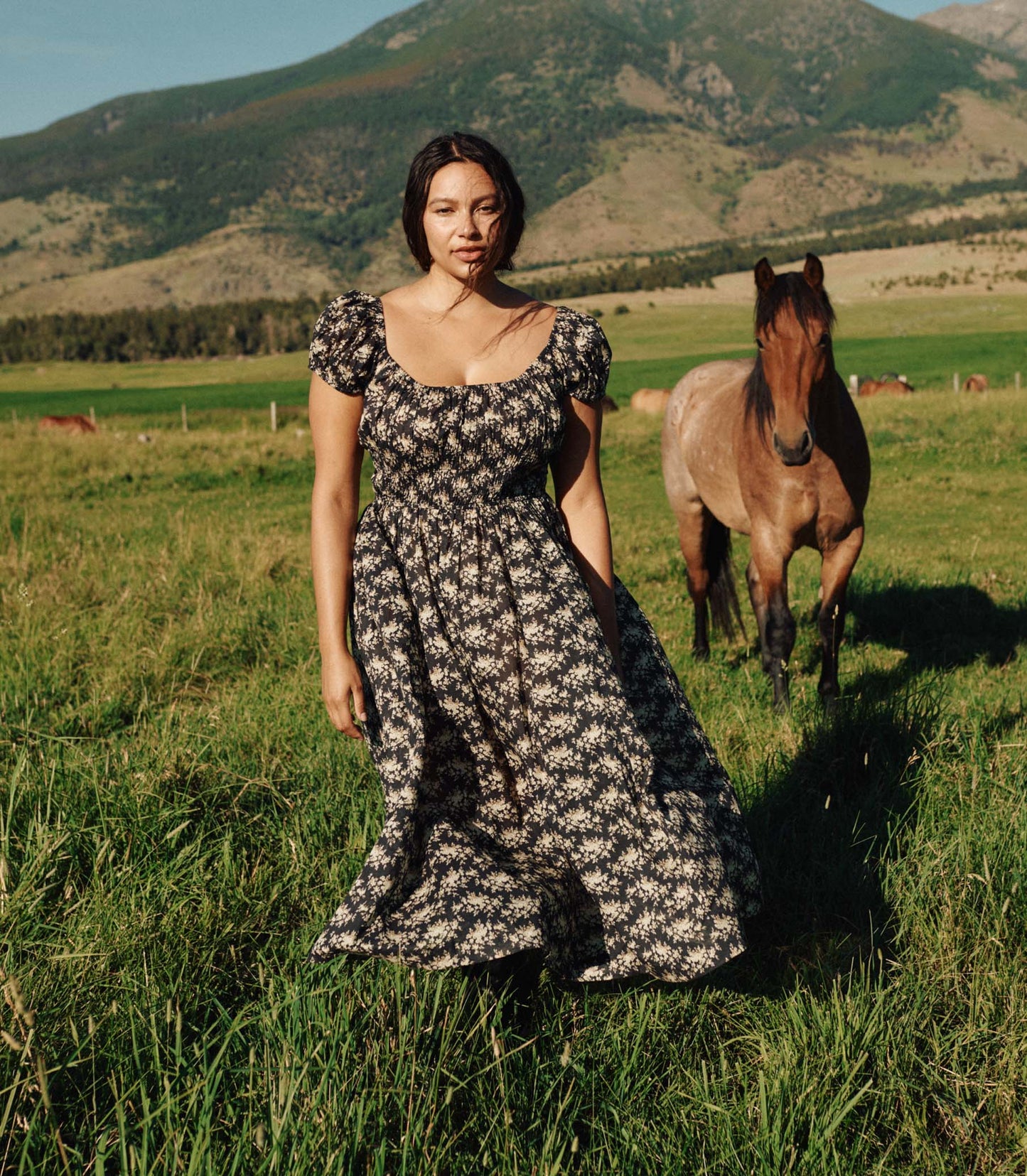Woman in a floral dress standing in a field with a horse and mountains in the background