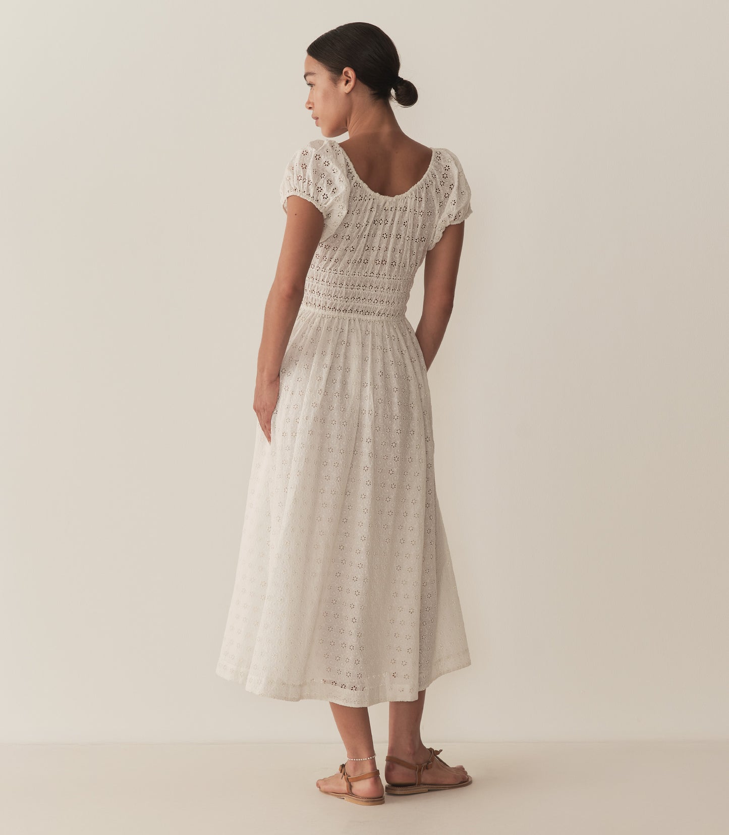 Zahara wears a long white eyelet dress with short puff sleeves, paired with tan sandals.