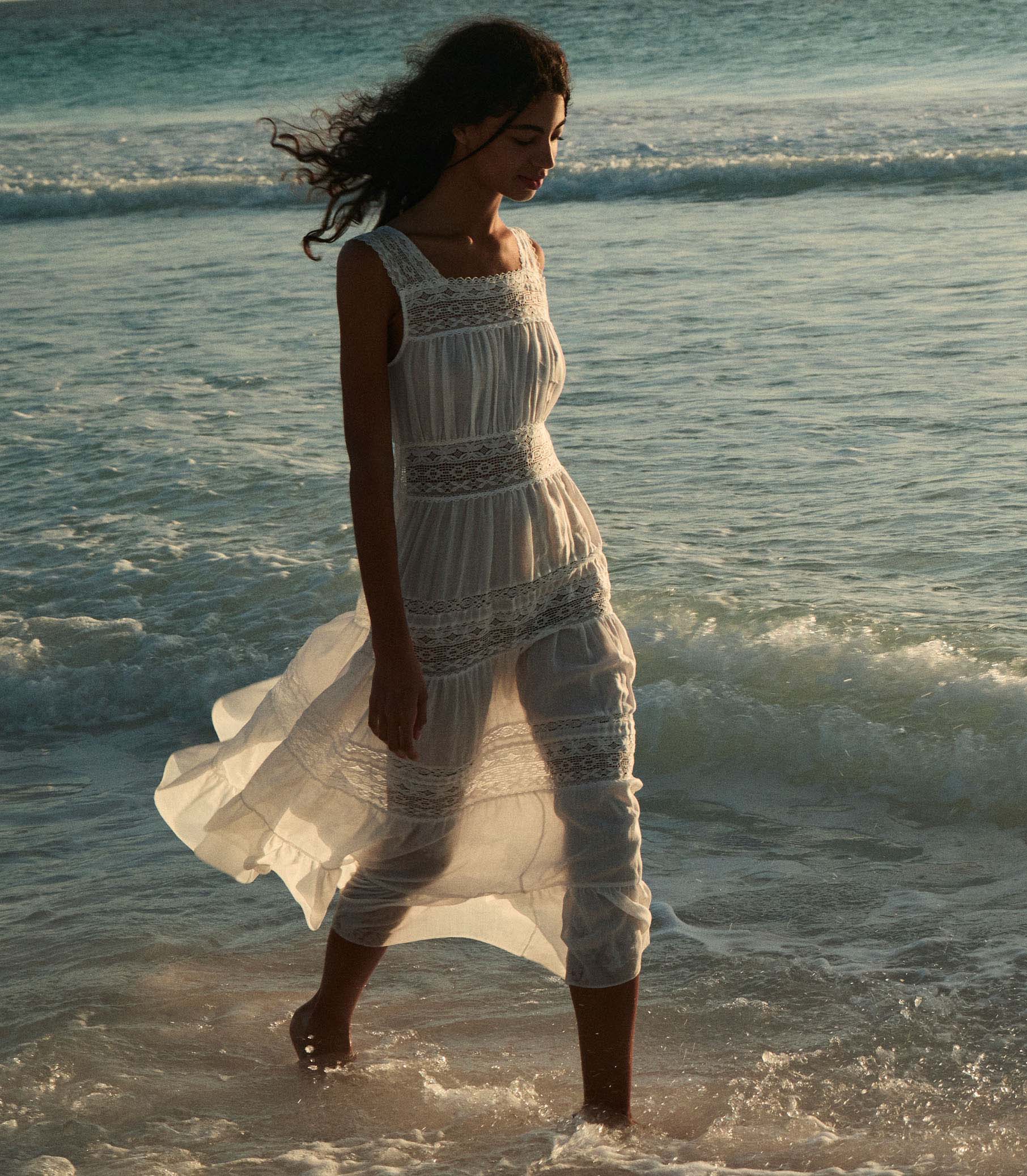 Rocio is wearing a long white sleeveless dress as she walks through the beach.