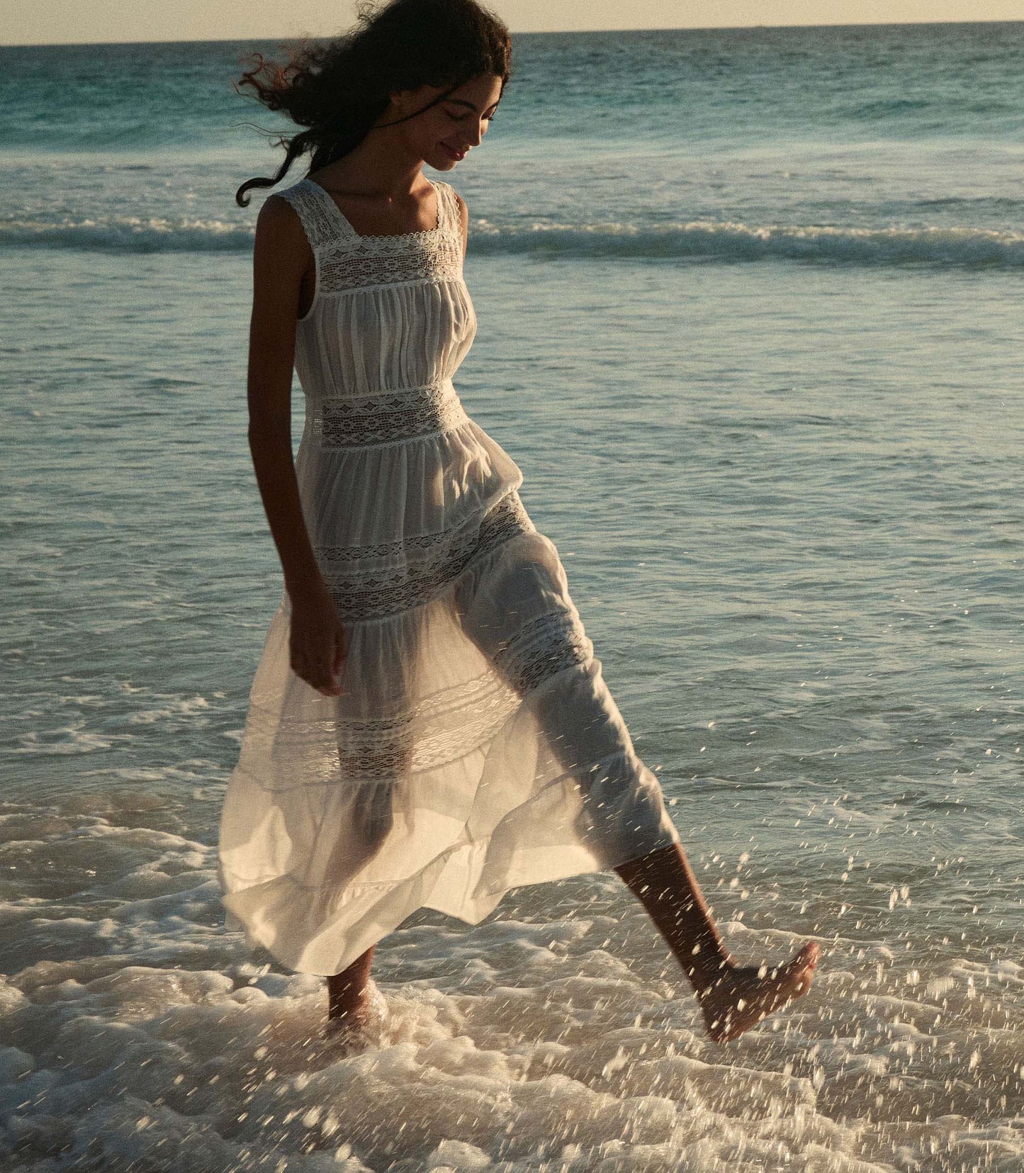 Rocio is wearing a long white sleeveless dress as she walks through the beach.