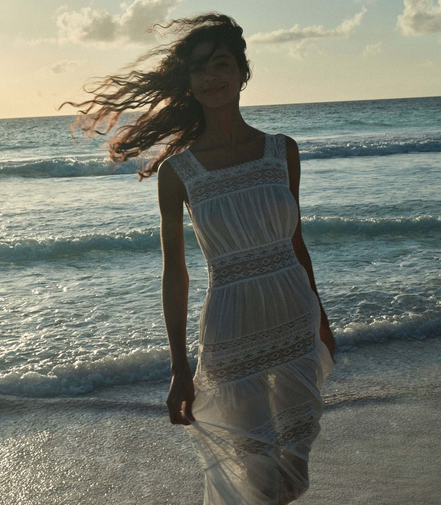 Rocio is wearing a long white sleeveless dress as she walks through the beach.