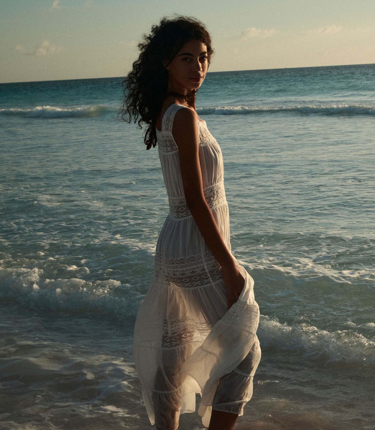 Rocio is wearing a long white sleeveless dress as she walks through the beach.