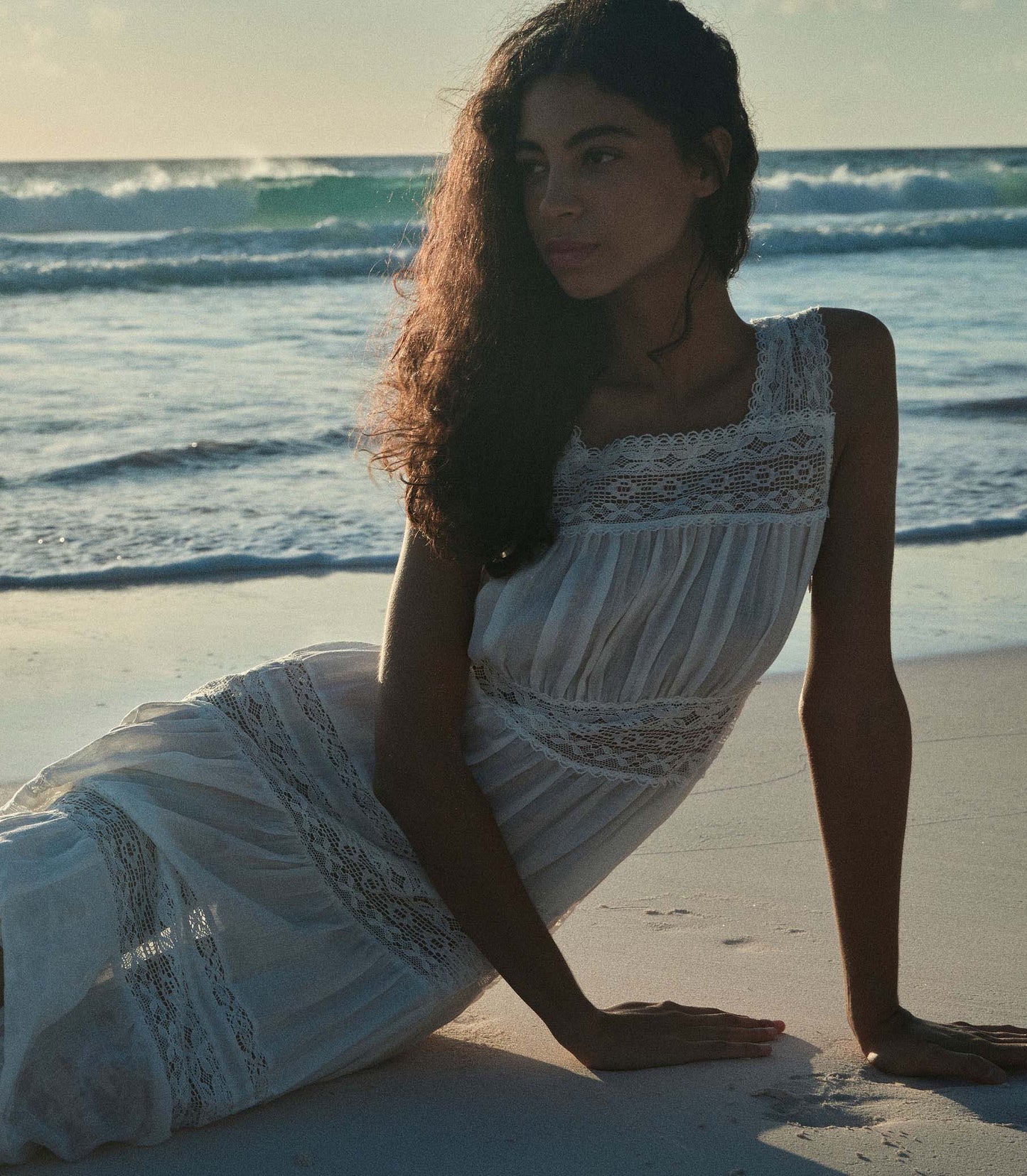 Rocio is wearing a long white sleeveless dress as she walks through the beach.