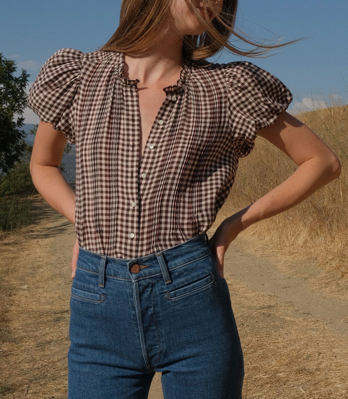 Woman wearing a checkered blouse and blue jeans standing outdoors with a scenic background.