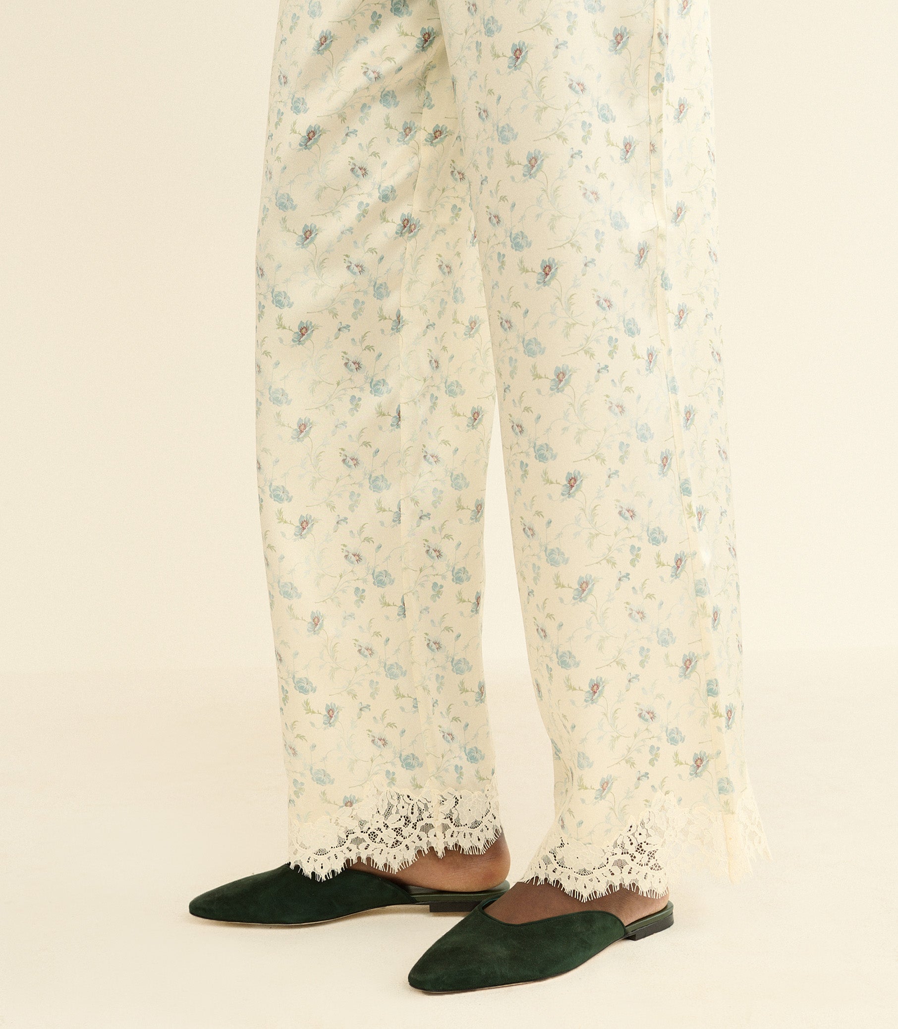 A close-up of a person's legs wearing cream-colored wide-leg trousers with a delicate blue and green floral print and lace trim, paired with dark green pointed-toe flats on a light beige background. 1
