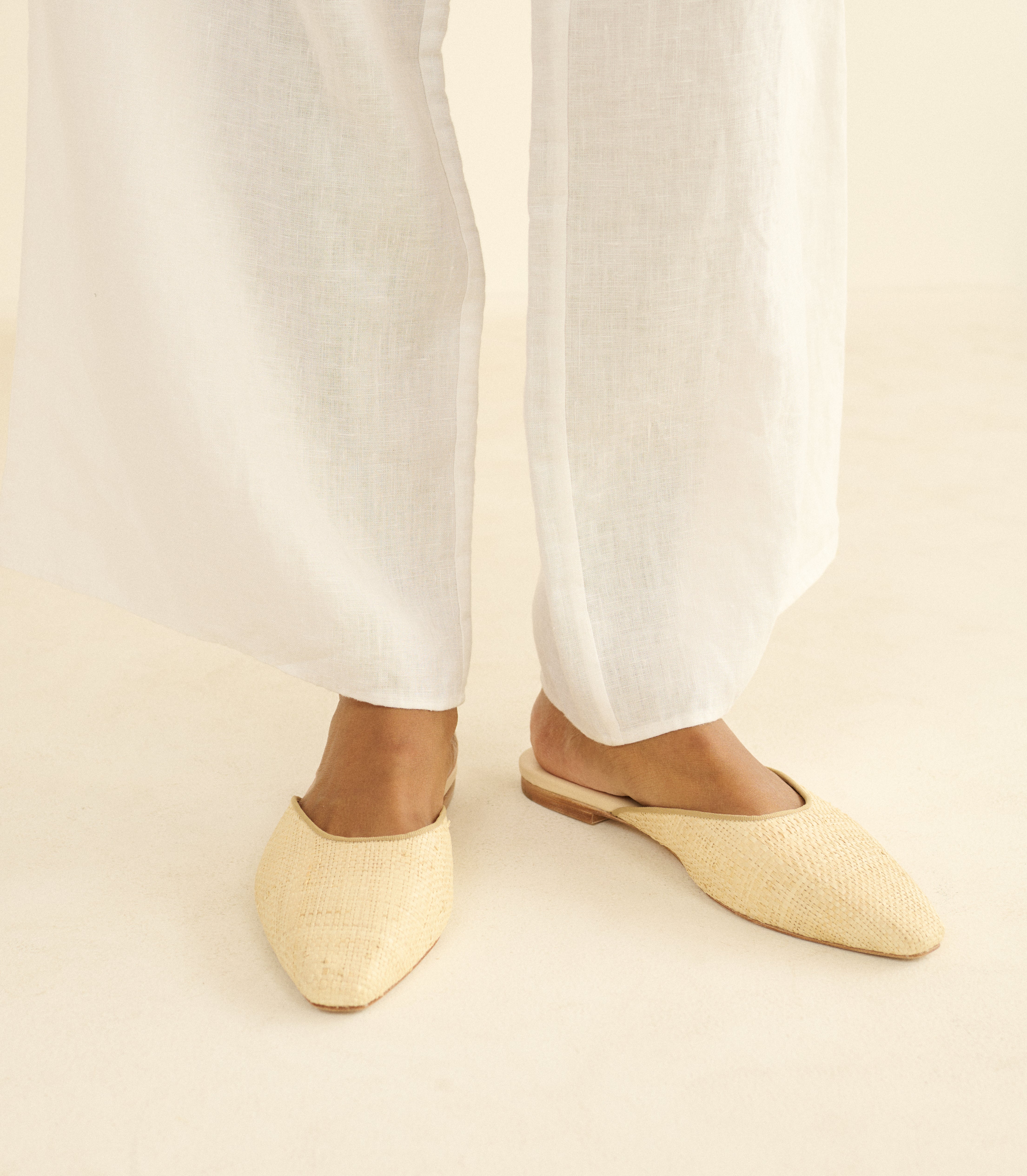 Person wearing beige woven flats with white pants on a neutral background
