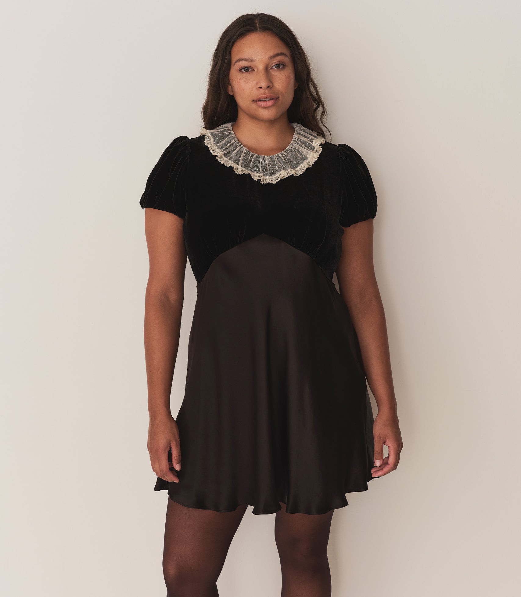 A woman wearing the Adelie Dress in Black, with a ruffled neckline, velvet bodice, and satin skirt.