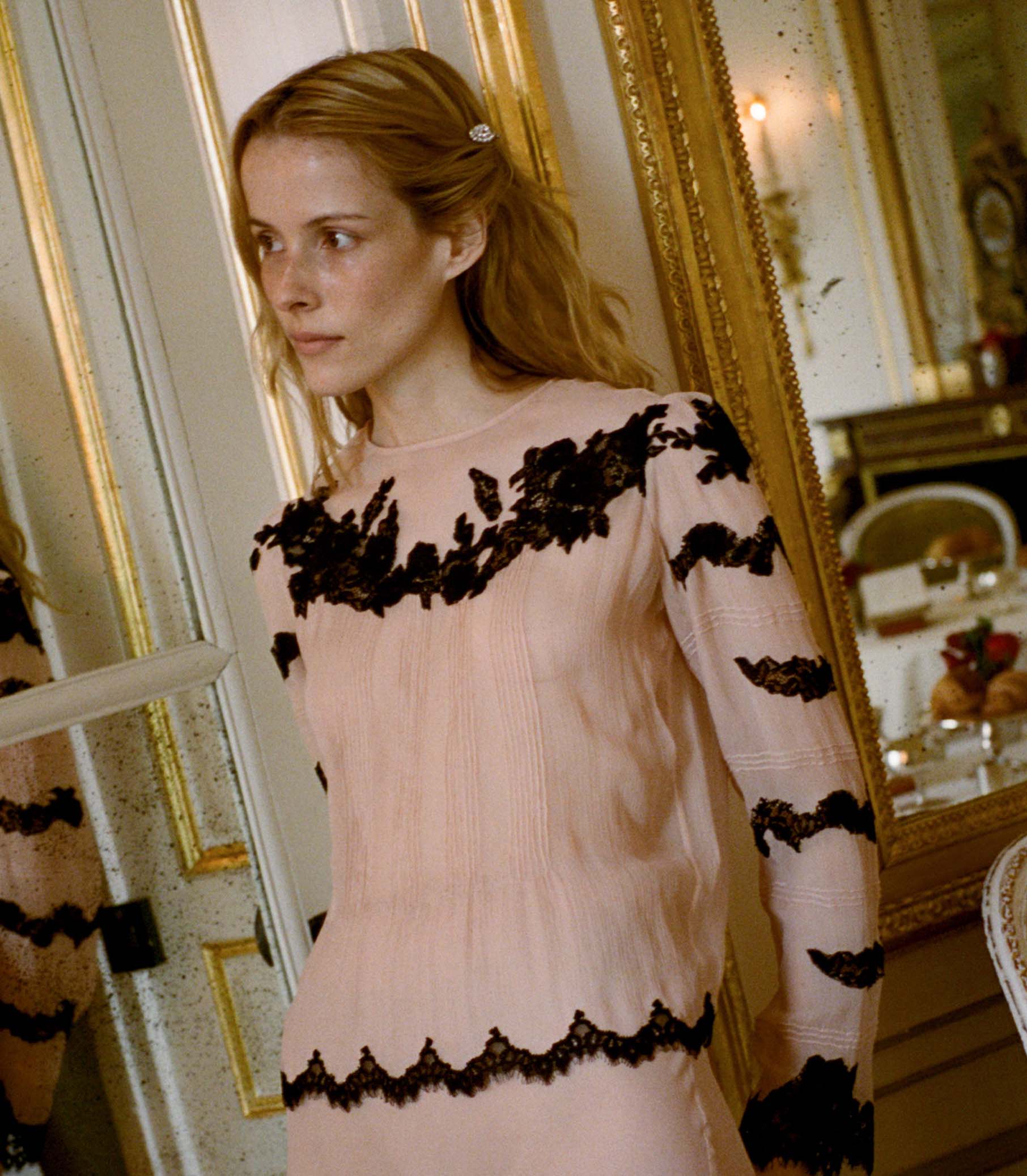 Woman wearing a pink blouse with black lace details in an elegant room.