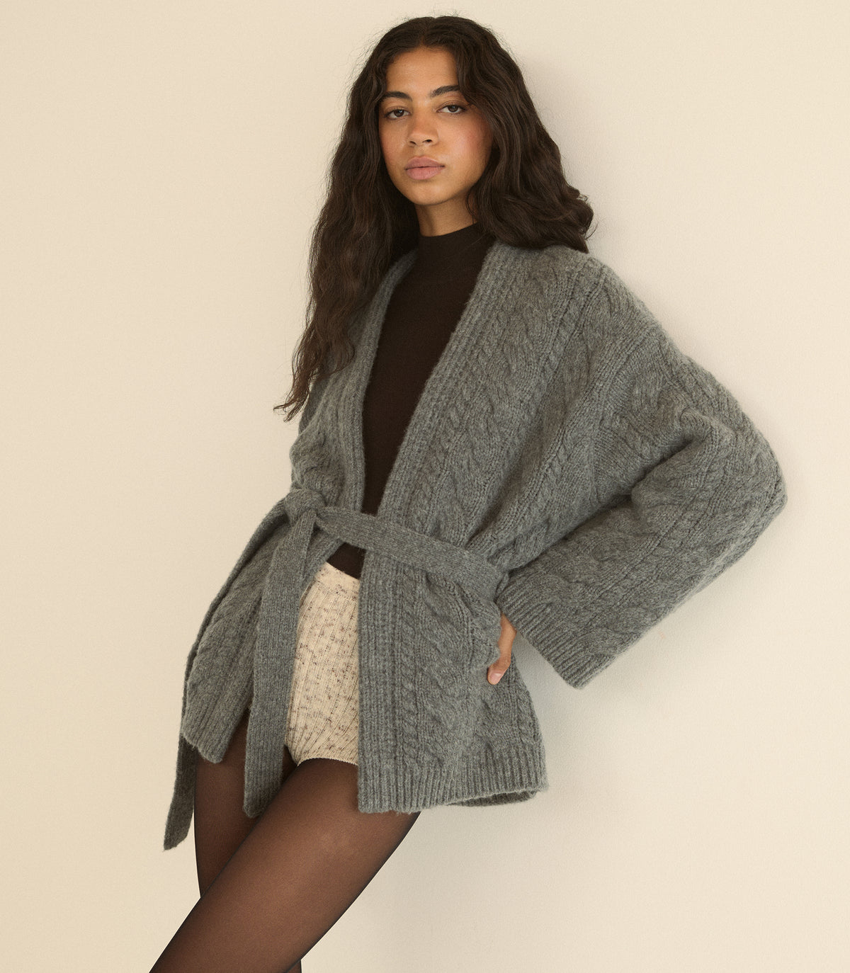 A woman wearing a chunky gray cable-knit cardigan with a tie waist, styled over a dark brown turtleneck and cream knit shorts, posing against a plain beige wall. 4
