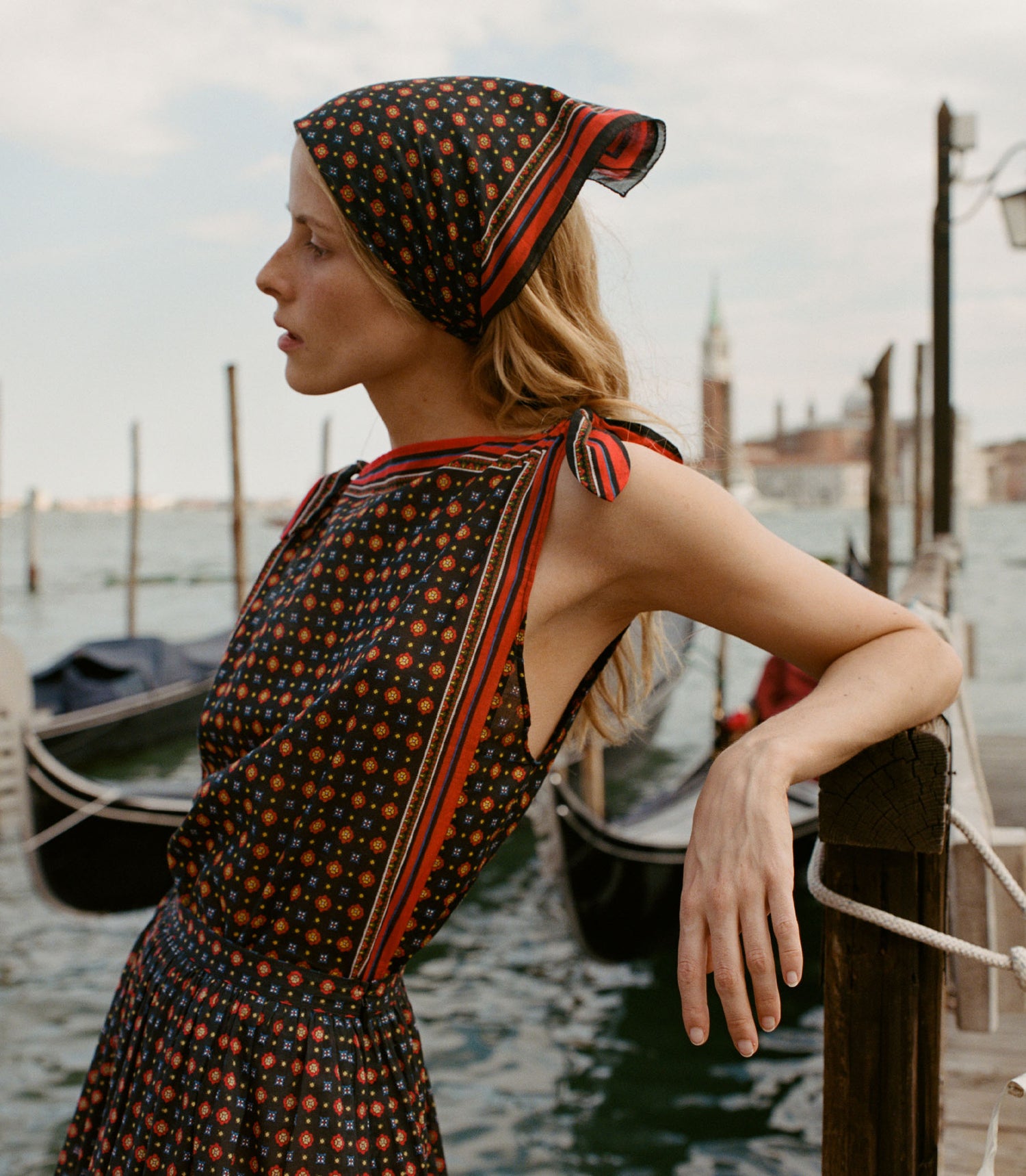 Woman in a patterned dress and headscarf by a dock with gondolas.