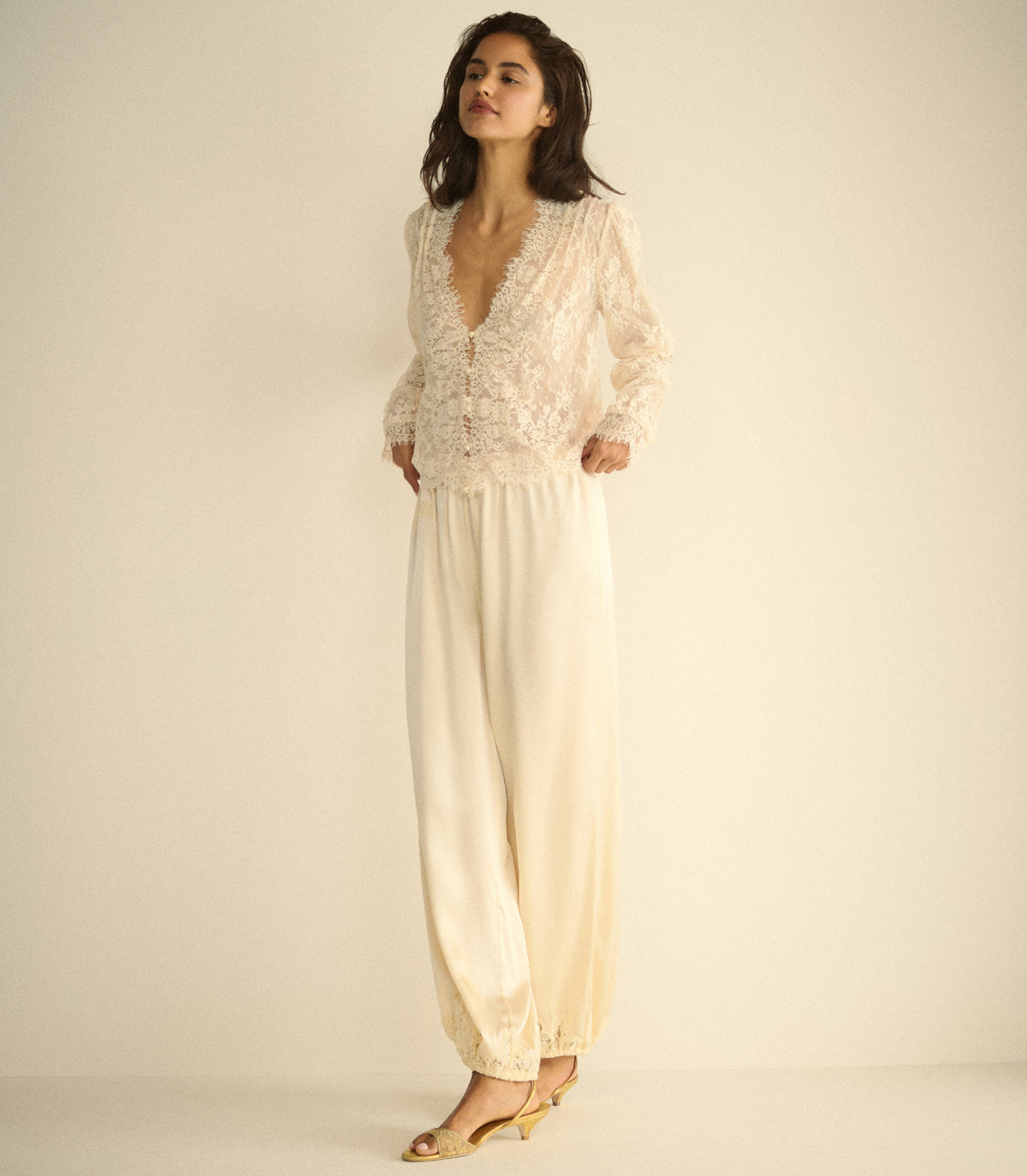 A woman wearing a sheer white lace blouse with long sleeves, paired with white silk pants and gold kitten heels, posed against a plain beige studio background.