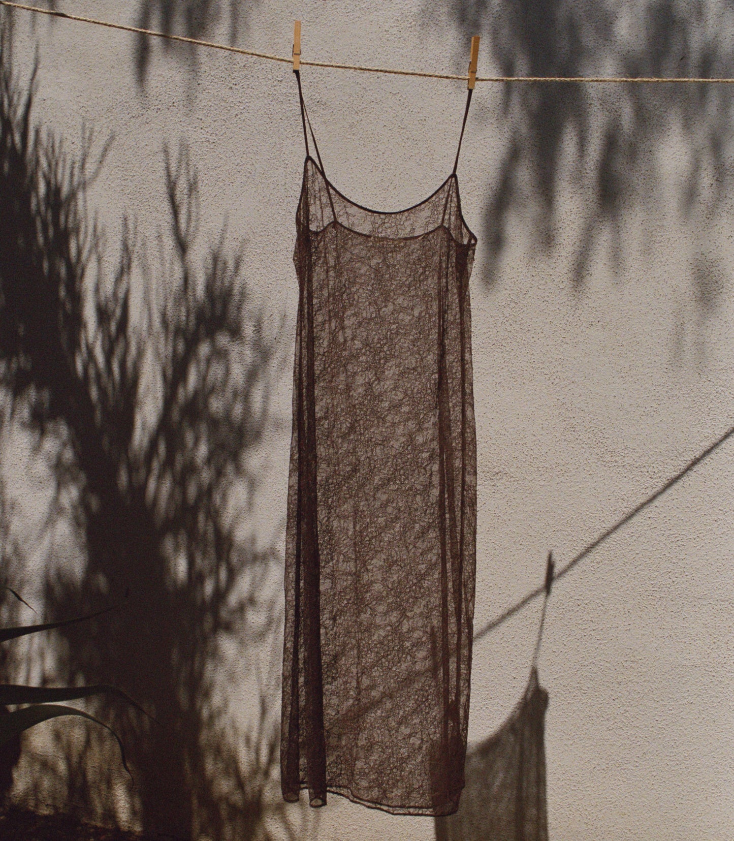 A sheer, dark brown lace slip dress hanging on a clothesline against a textured wall with tree shadows cast across it.