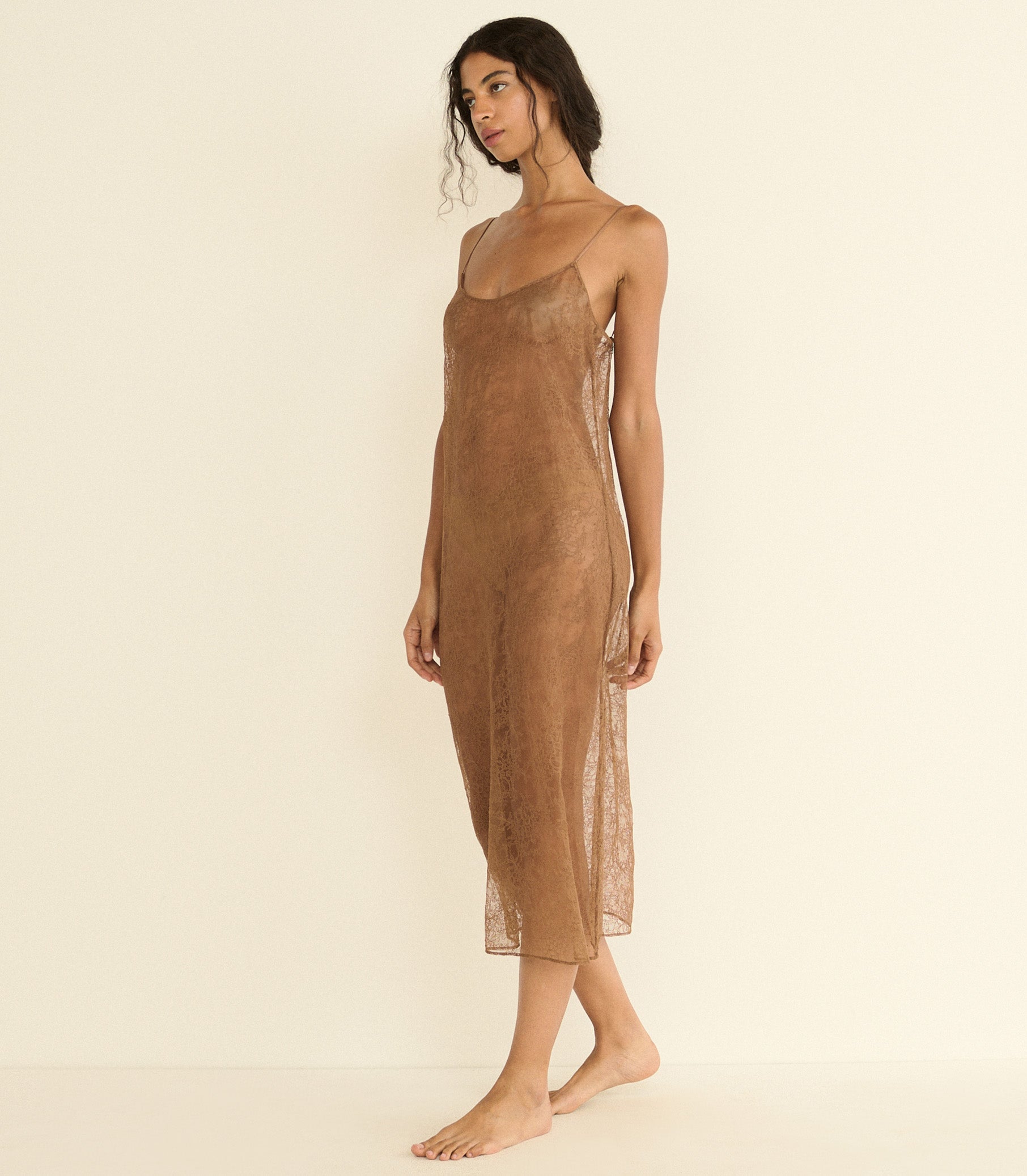 A woman modeling a sheer, lace brown slip dress with spaghetti straps against a plain, warm off-white studio background. 1
