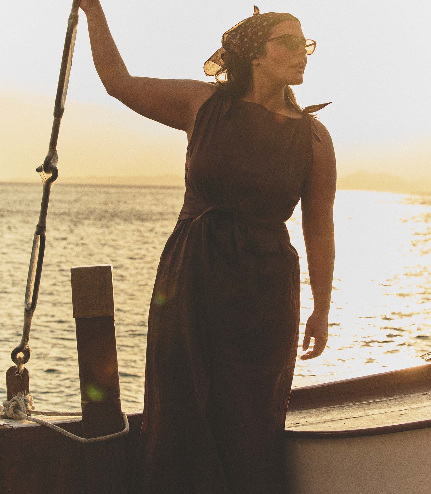 Woman in a brown dress standing on a boat with a sunset or sunrise in the background.