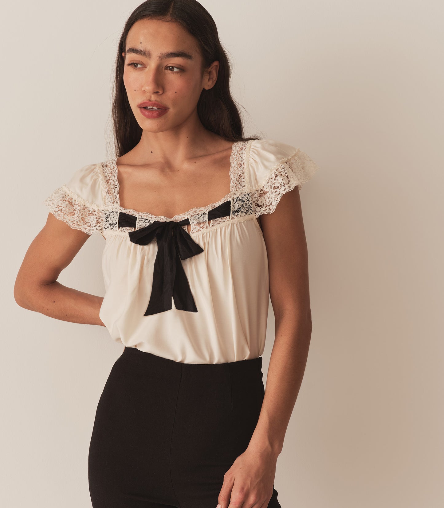 Zahara wears the  Anaya Top, which features a breezy silhouette, delicate flutter sleeves, and a soft V-neckline trimmed with intricate lace and threaded with a contrast silk ribbon.