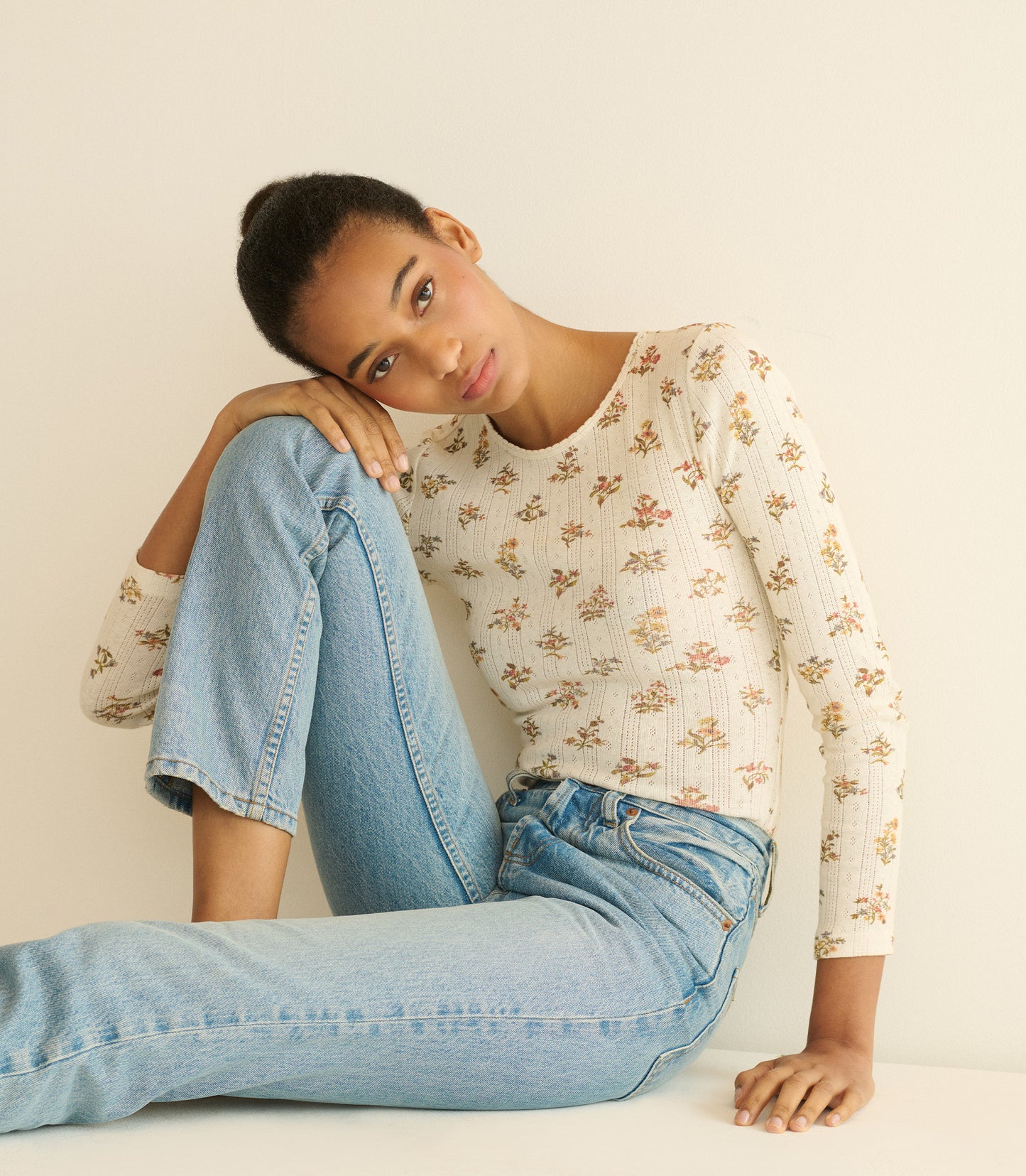 Woman wearing a floral top and blue jeans sitting on a white surface.