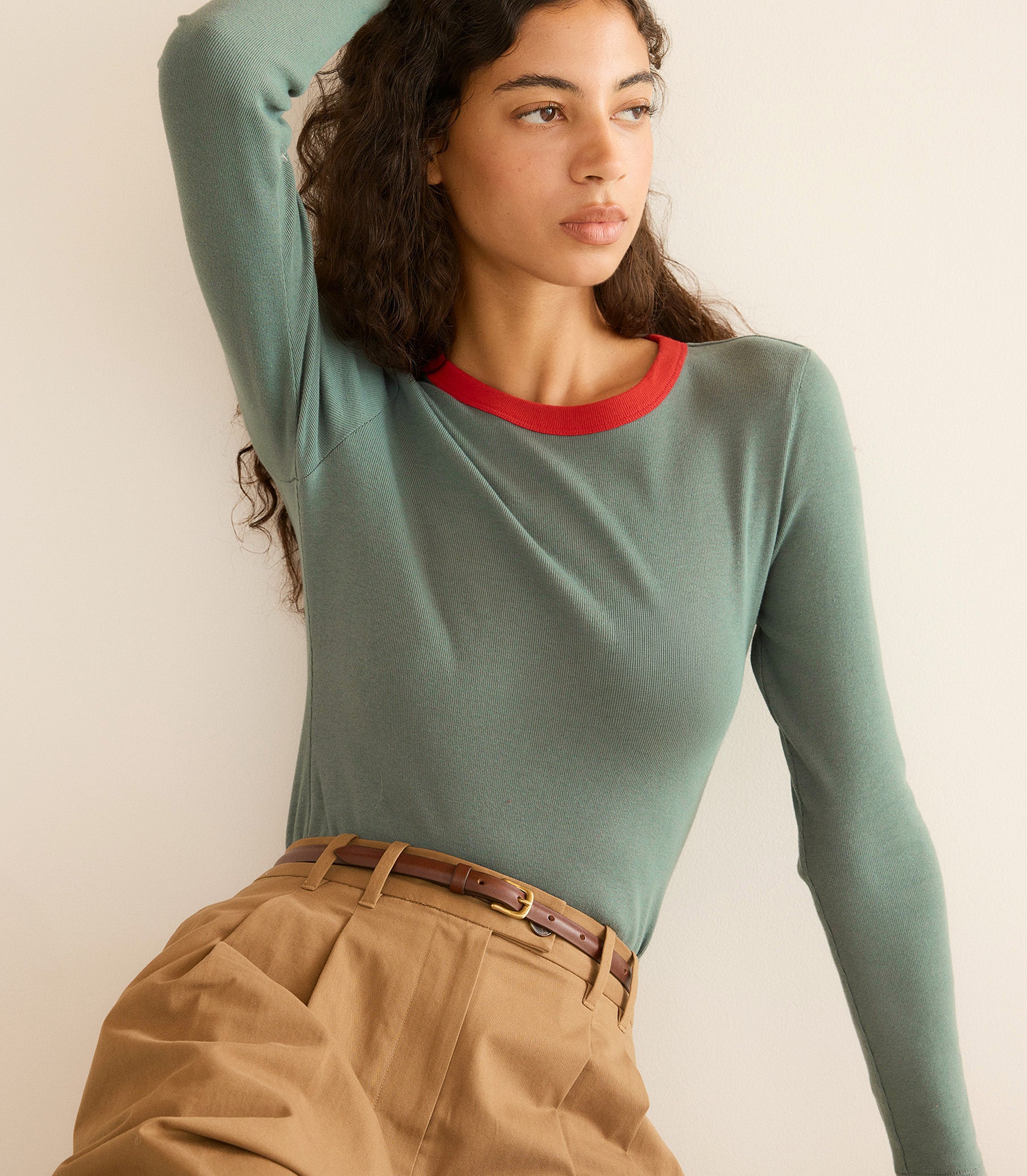 Woman wearing a green long-sleeve top with red neckline and beige pants against a plain background