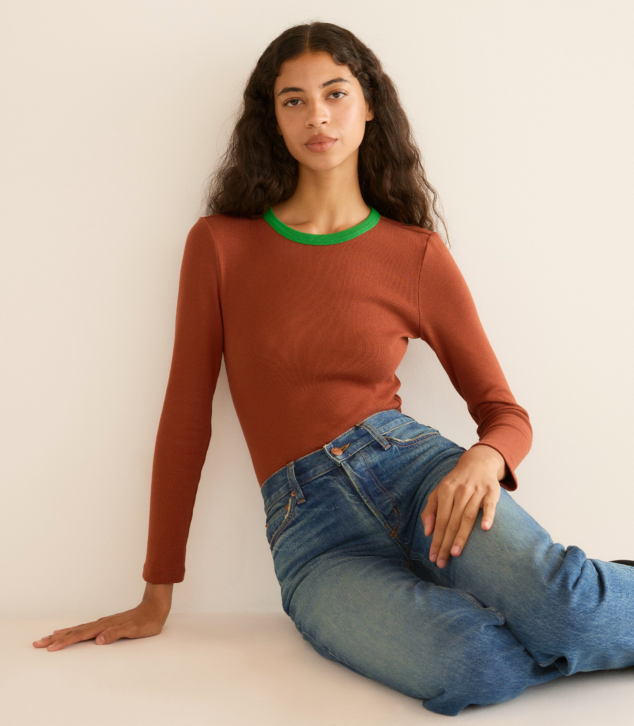 Woman wearing a brown sweater with green trim and blue jeans sitting against a white wall.