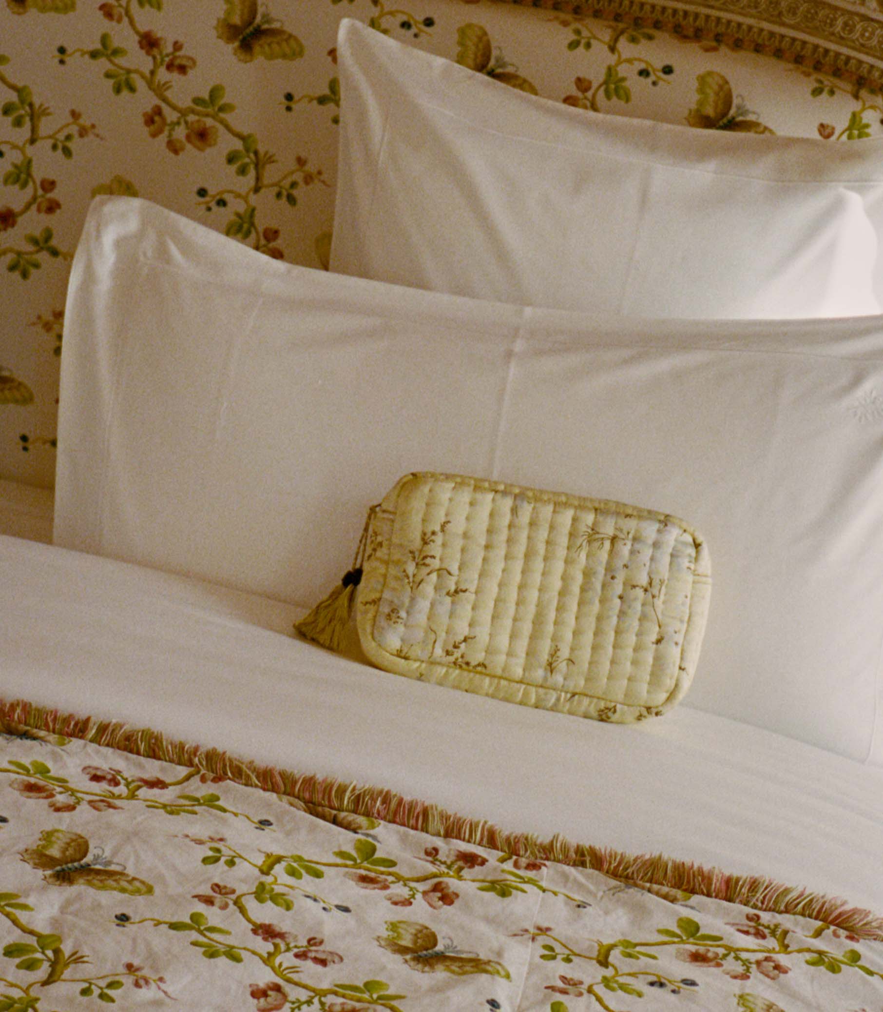 Light yellow floral pouch against white pillows on a bed with floral bedding and wallpaper.