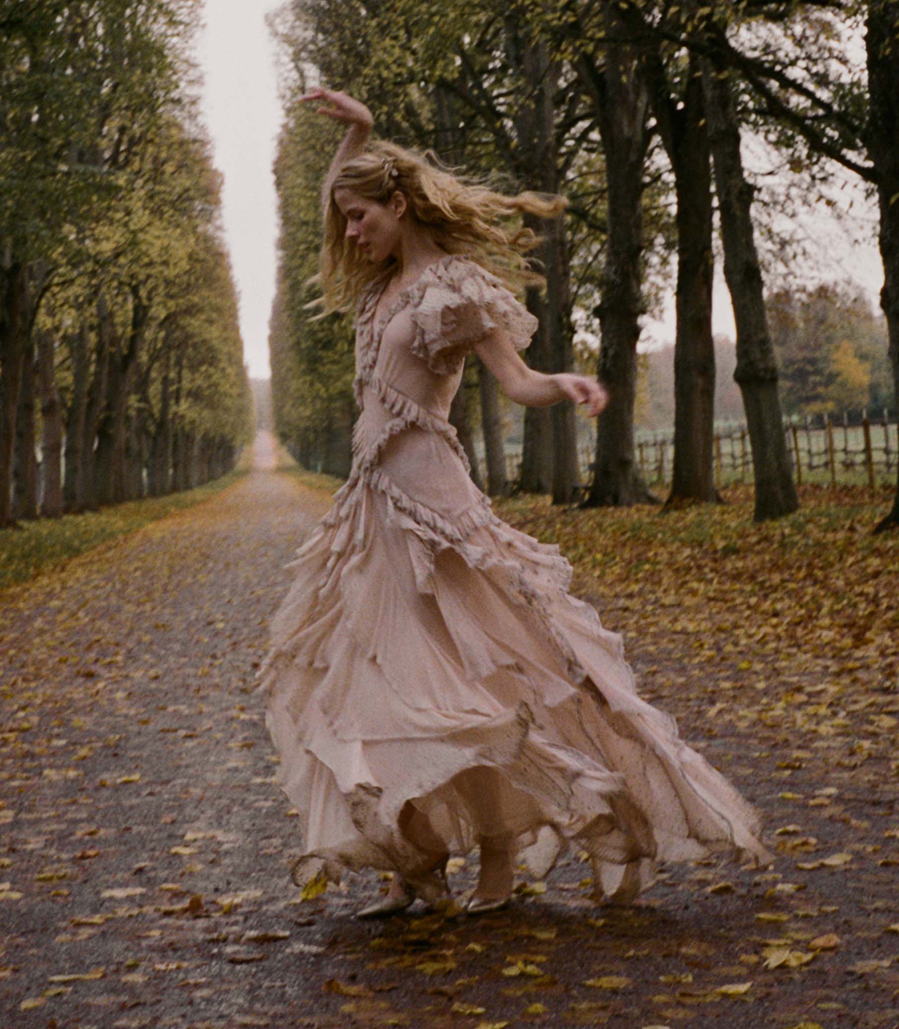 Woman in a long, antique blush dress dancing on a tree-lined path with fallen leaves. 2
