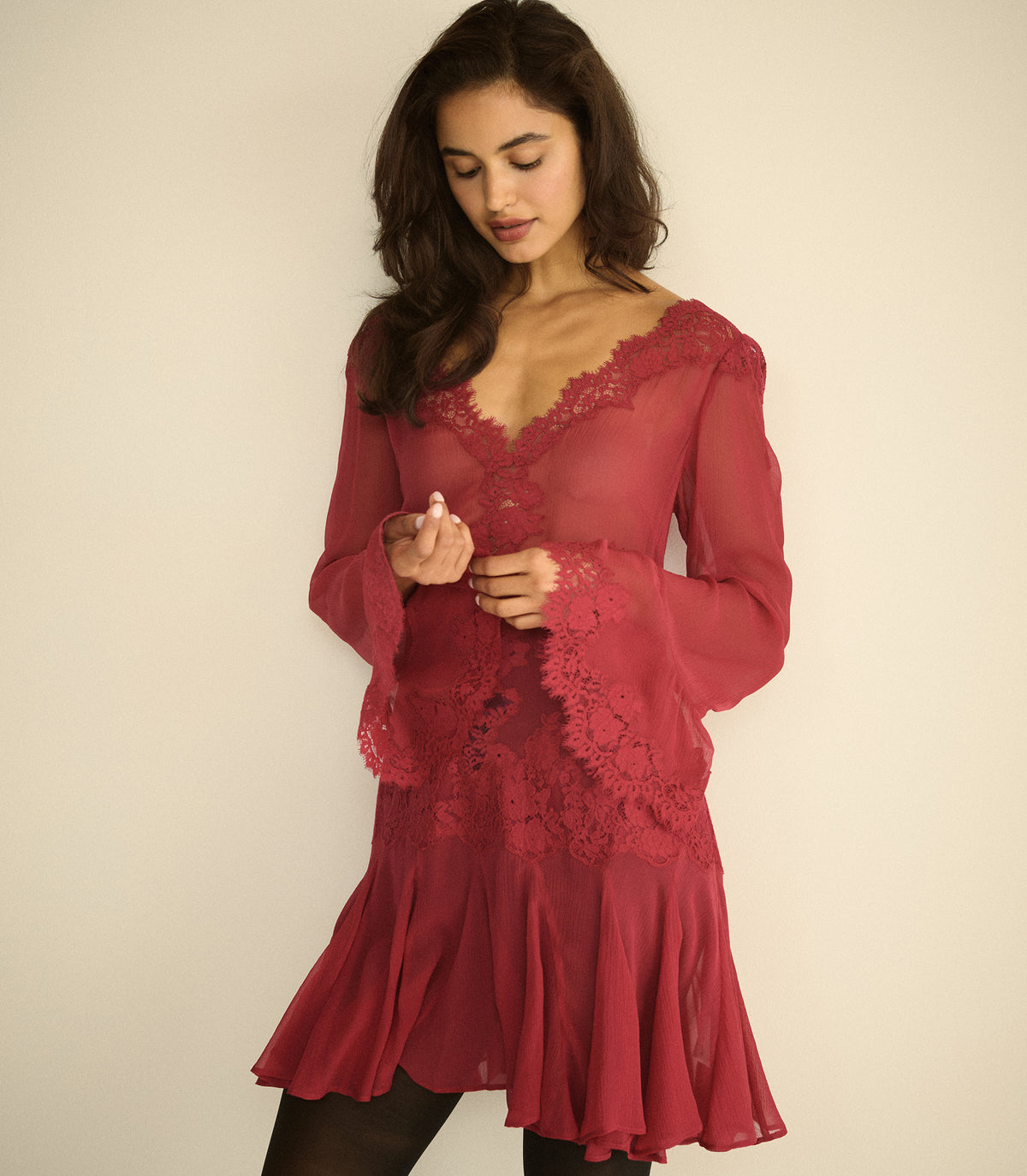A woman wearing a long-sleeved, deep red lace and mesh mini dress with a ruffled hem, standing against a plain beige background.