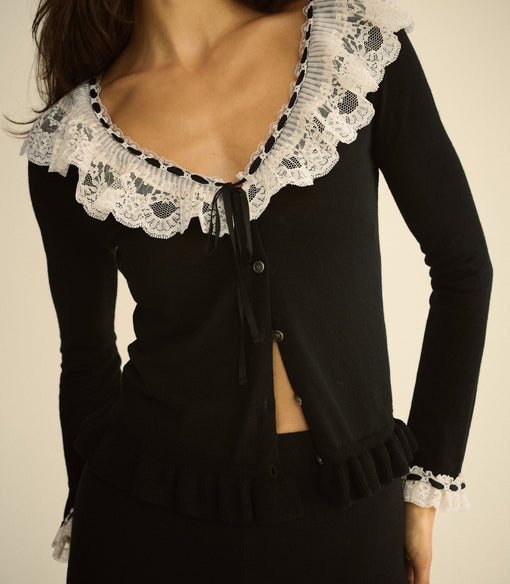 A close-up of a model wearing a black long-sleeve cardigan with a large white lace ruffle collar and matching lace cuffs, set against a plain beige background.