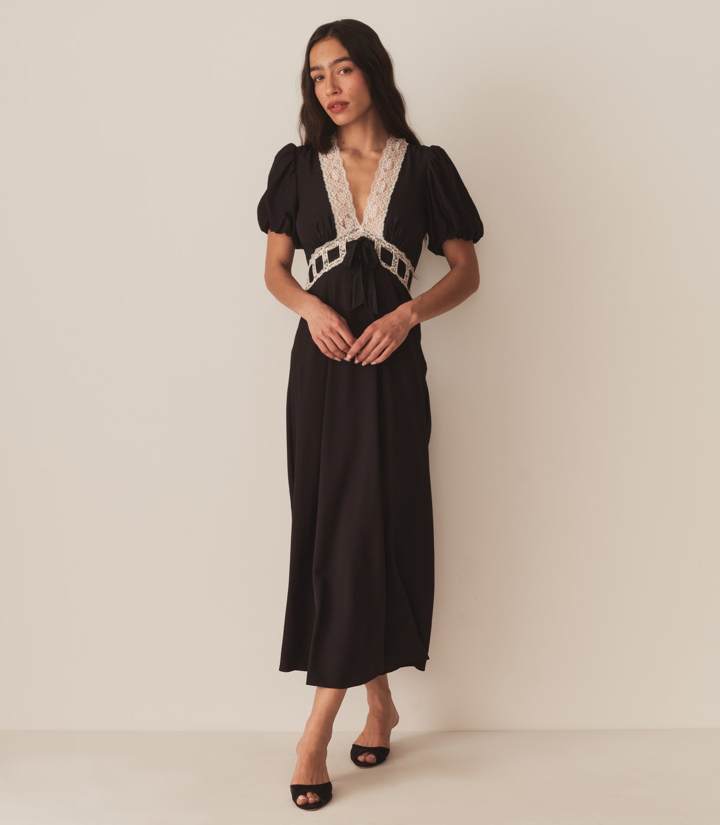 Zahara wears the Augustina Dress in Black, which features short puffed sleeves and a deep V-neckline trimmed with intricate lace.