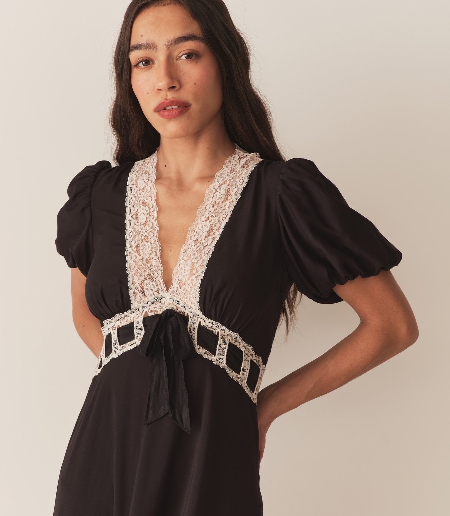 Zahara wears the Augustina Dress in Black, which features short puffed sleeves and a deep V-neckline trimmed with intricate lace.