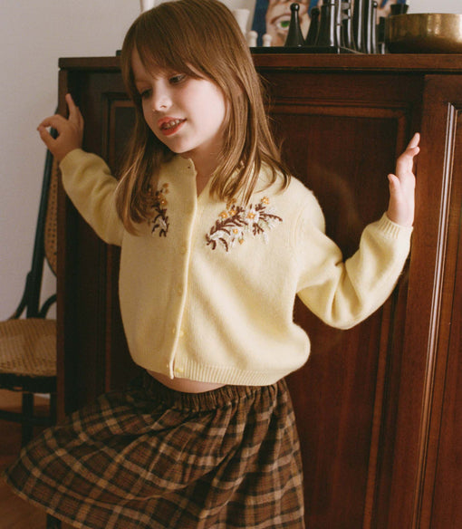 Young girl in a yellow cardigan with floral embroidery and plaid skirt standing in a room.