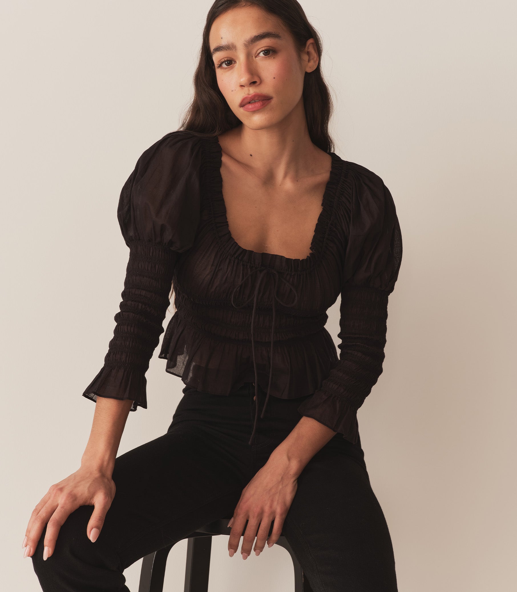 A woman wearing the Baptisia Top in Black, featuring puffed shoulders and ruching at the arms and bodice.