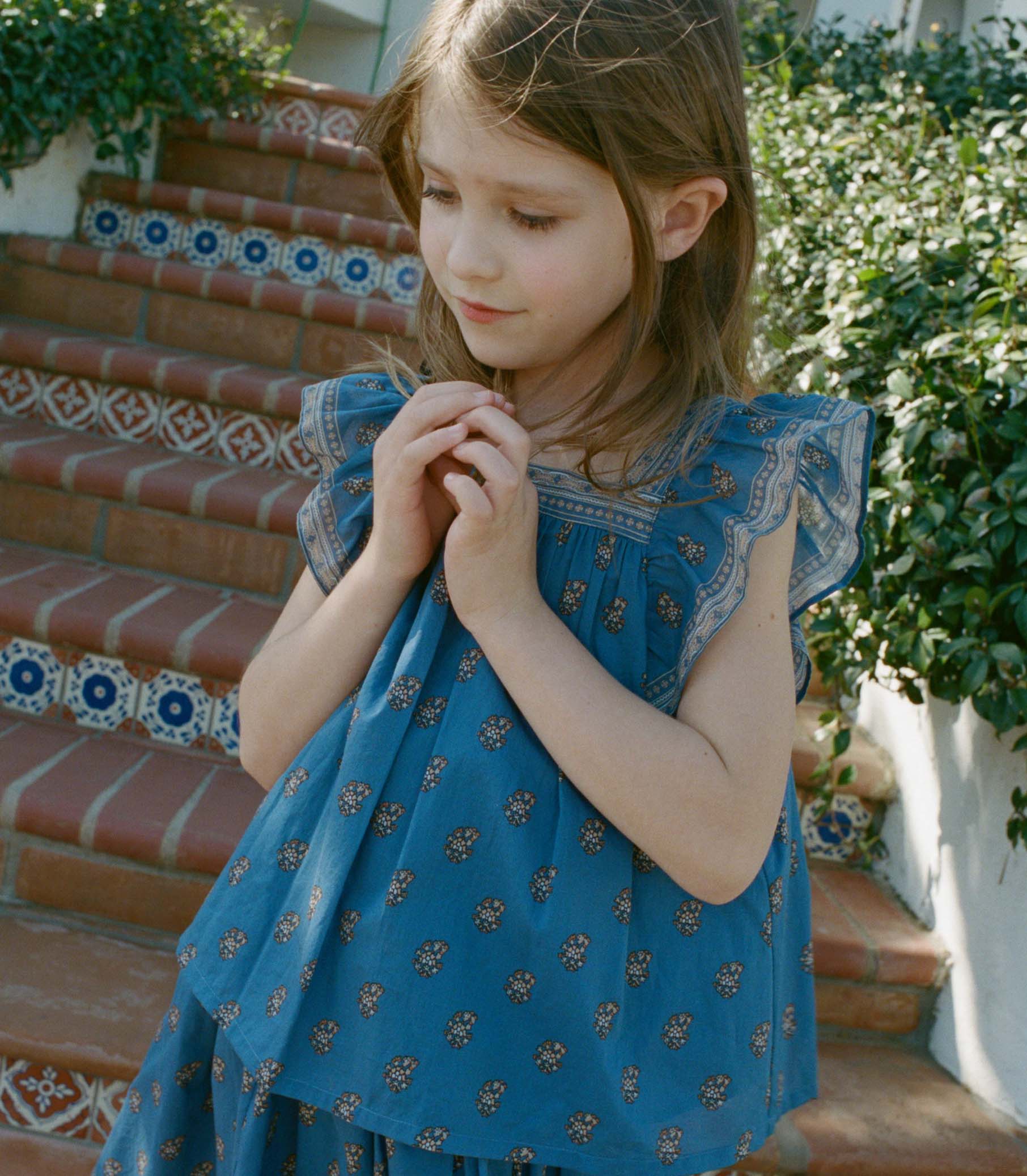 Young girl in a blue paisley top standing on outdoor steps with greenery. 1
