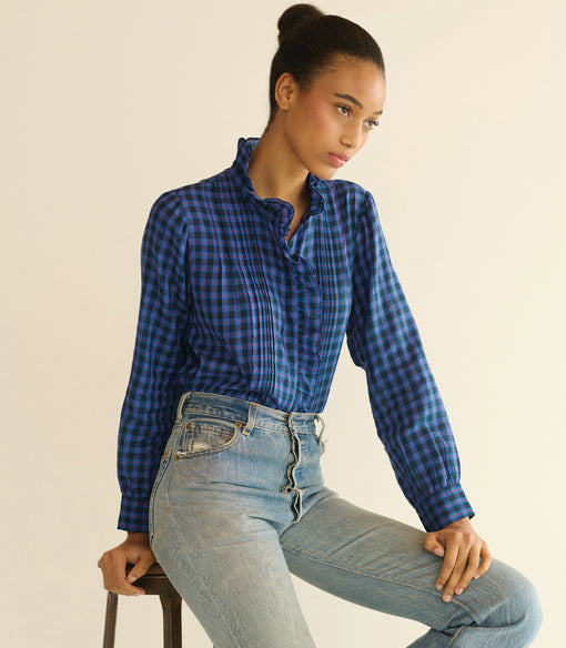 Woman wearing a blue checkered shirt and light blue jeans sitting on a stool against a beige background