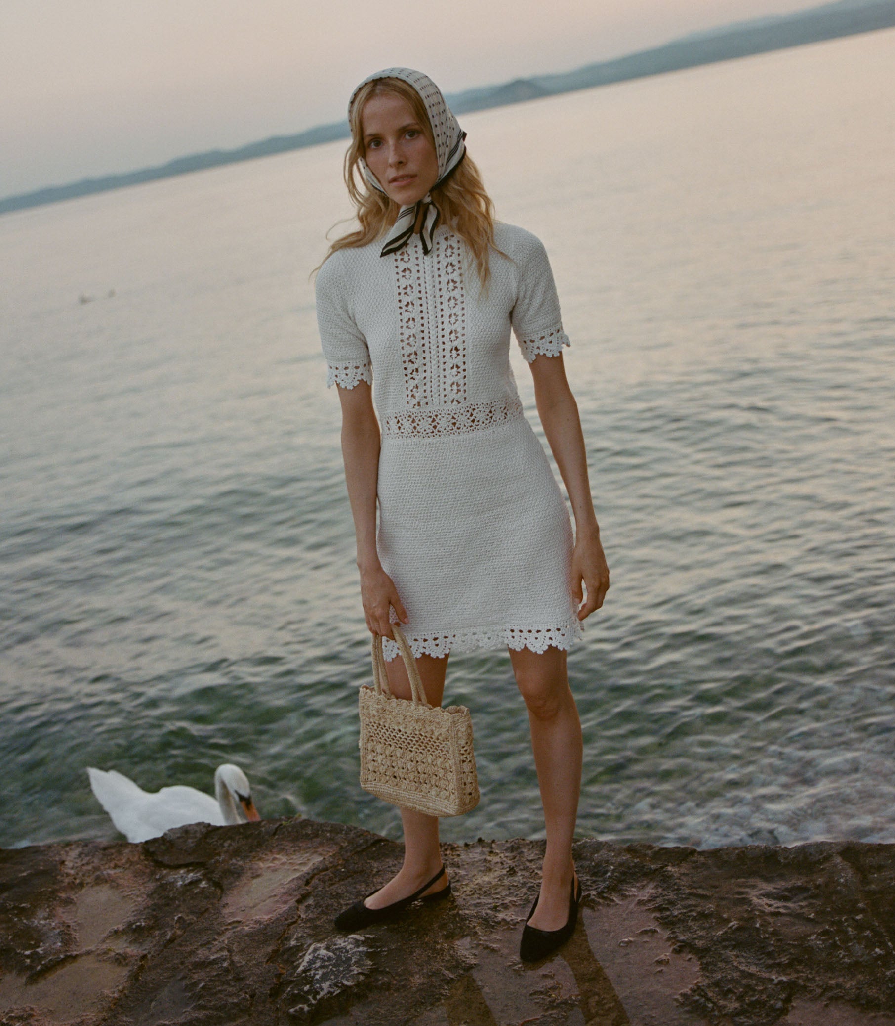 Woman in a white dress standing by a body of water with a swan in the background.