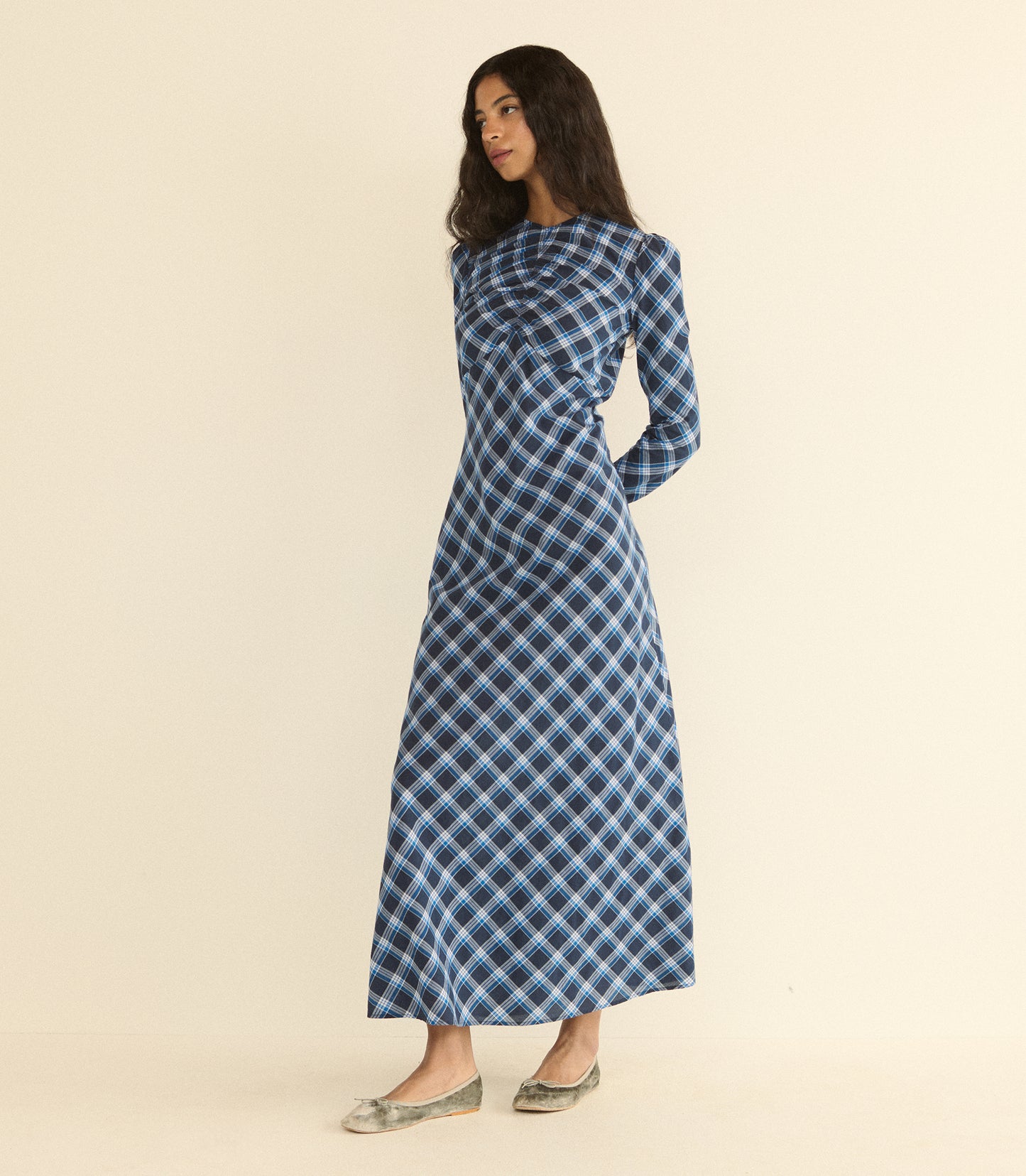 A woman modeling a long-sleeved, high-neck maxi dress with a blue, black, and white plaid pattern, standing against a solid beige studio background.