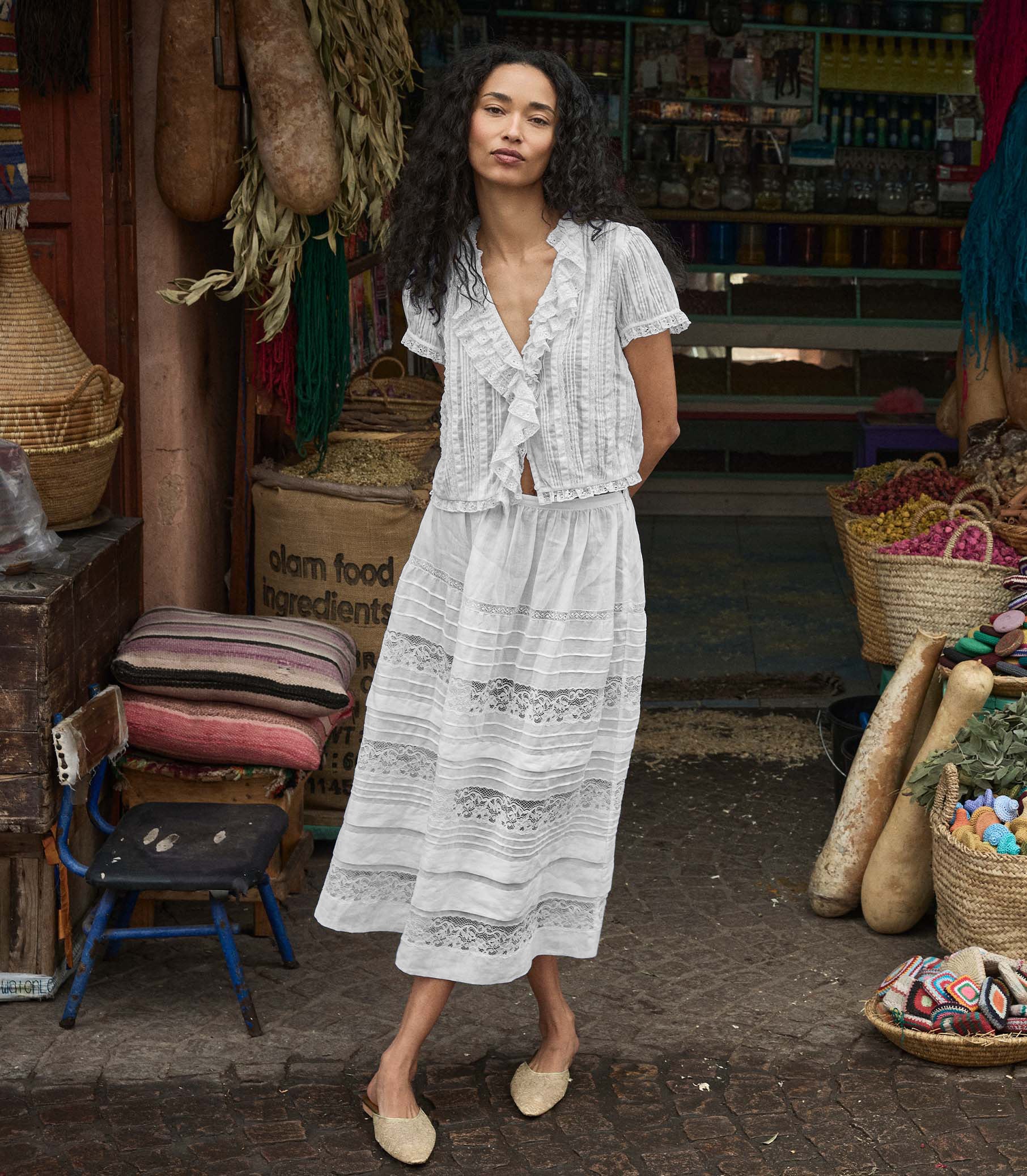 Woman in a white lace blouse and skirt standing in a market setting with various items around her. 1
