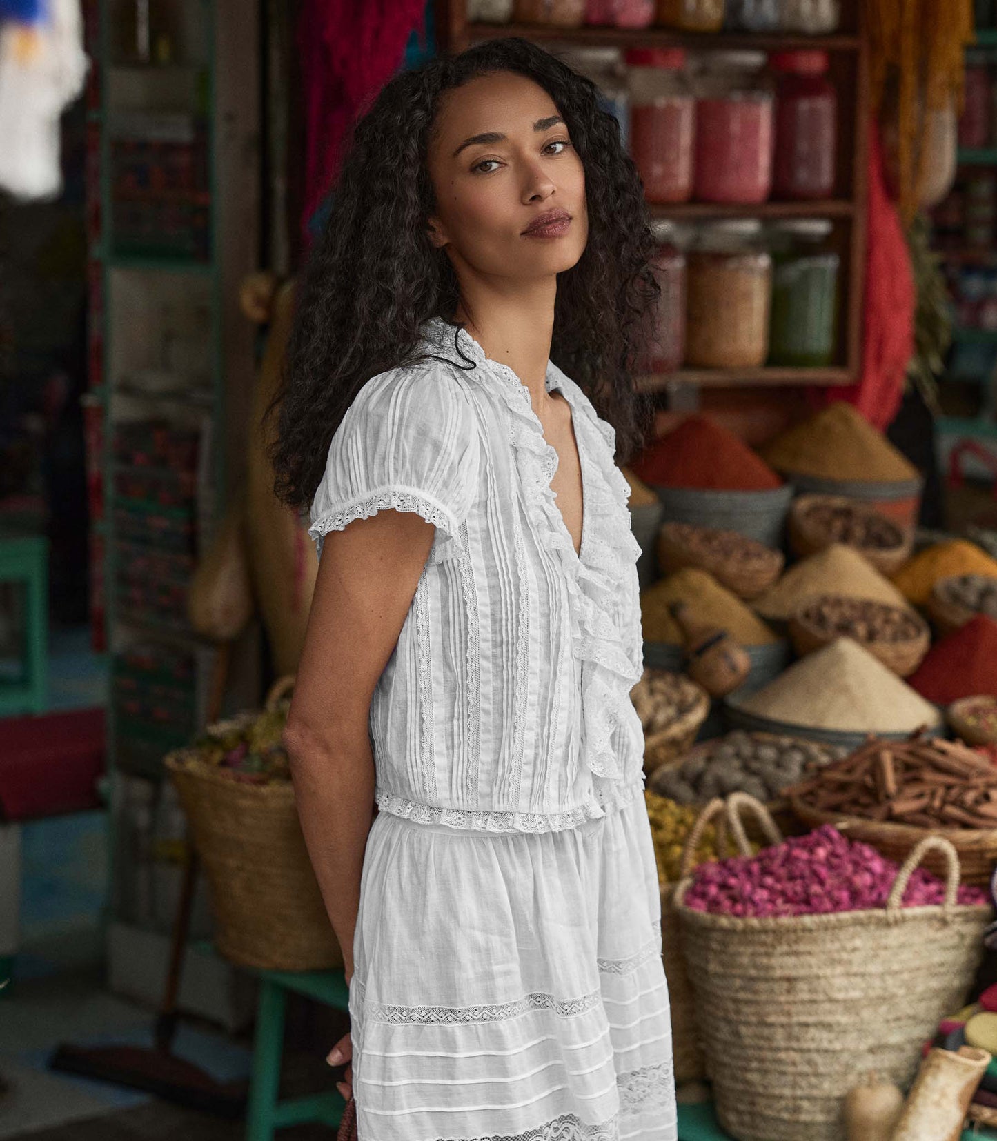 Woman in a white blouse standing in a market setting with various products and baskets.