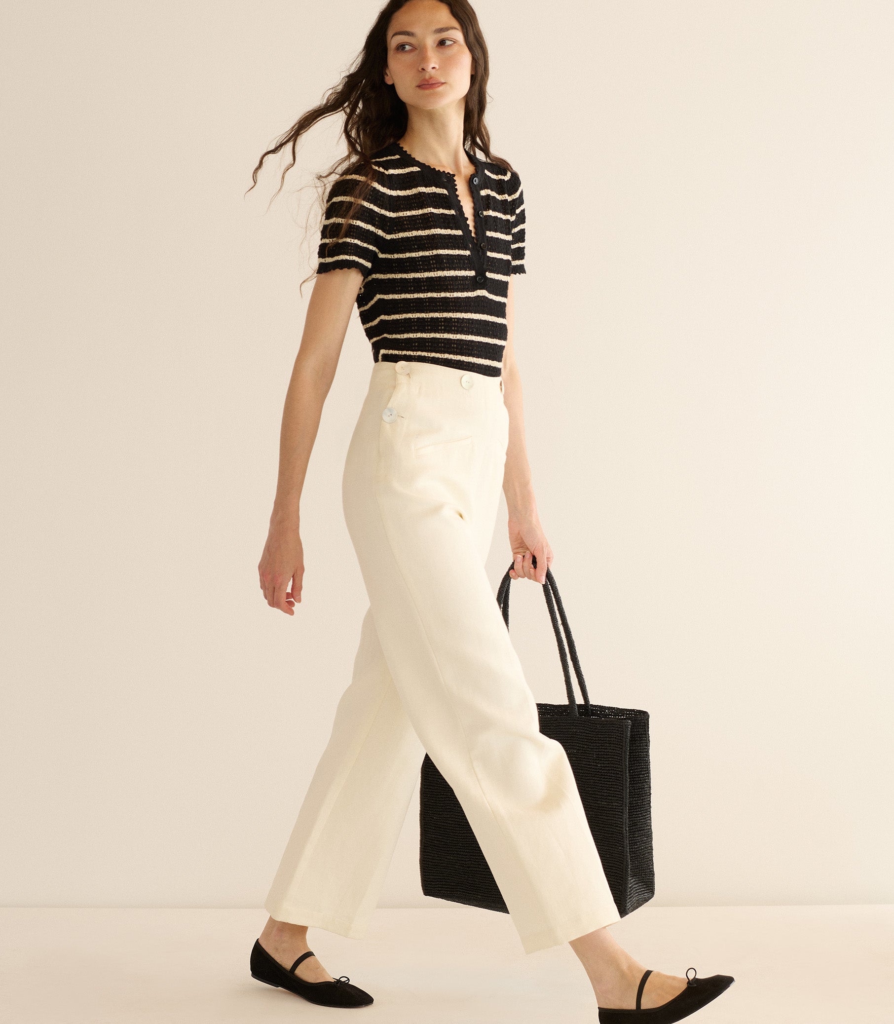 Woman wearing a black and white striped shirt and white pants, holding a black tote bag on a beige background